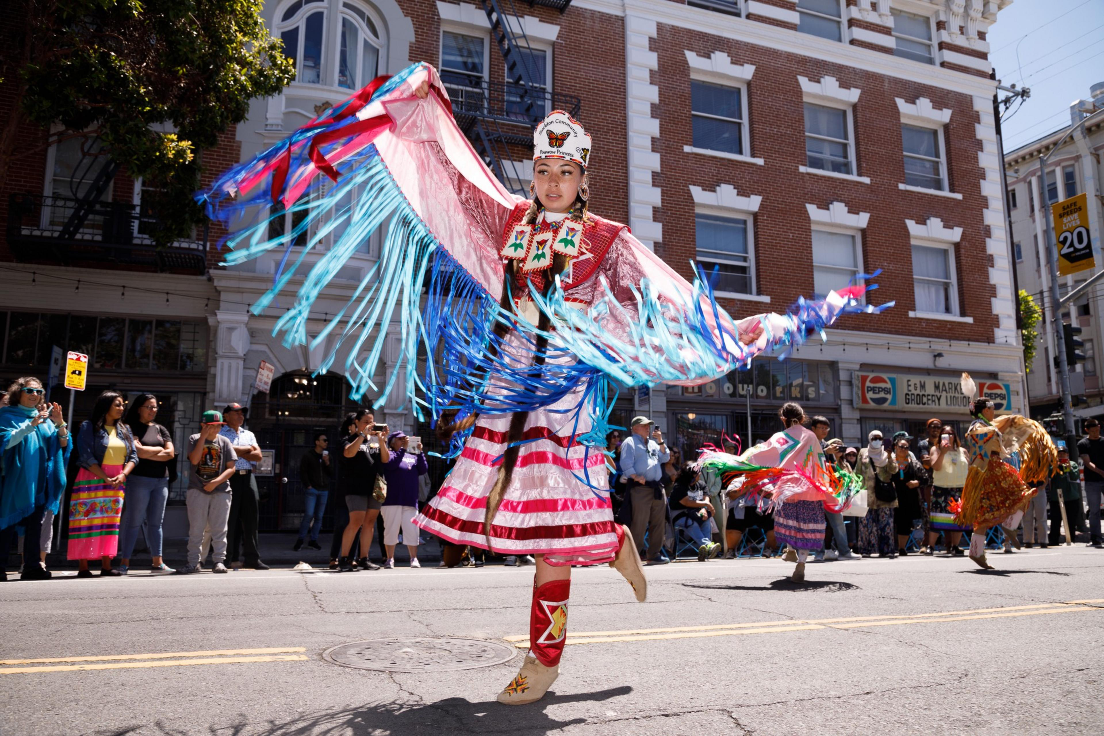 A dancer in vibrant traditional attire performs on the street, with spectators in the background.