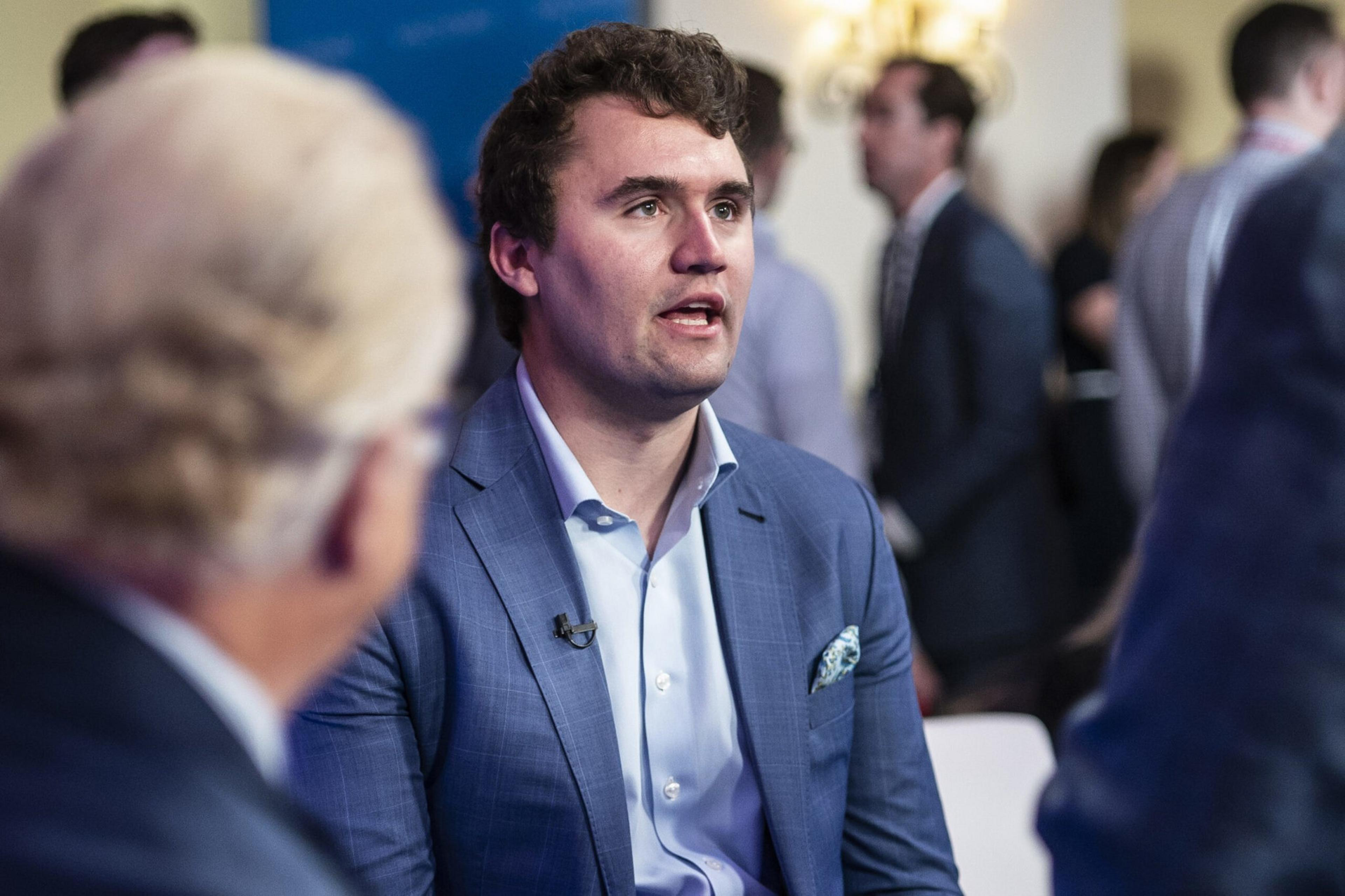 A man in a blue suit and light blue shirt speaks while seated among a group of people in a social or professional gathering.