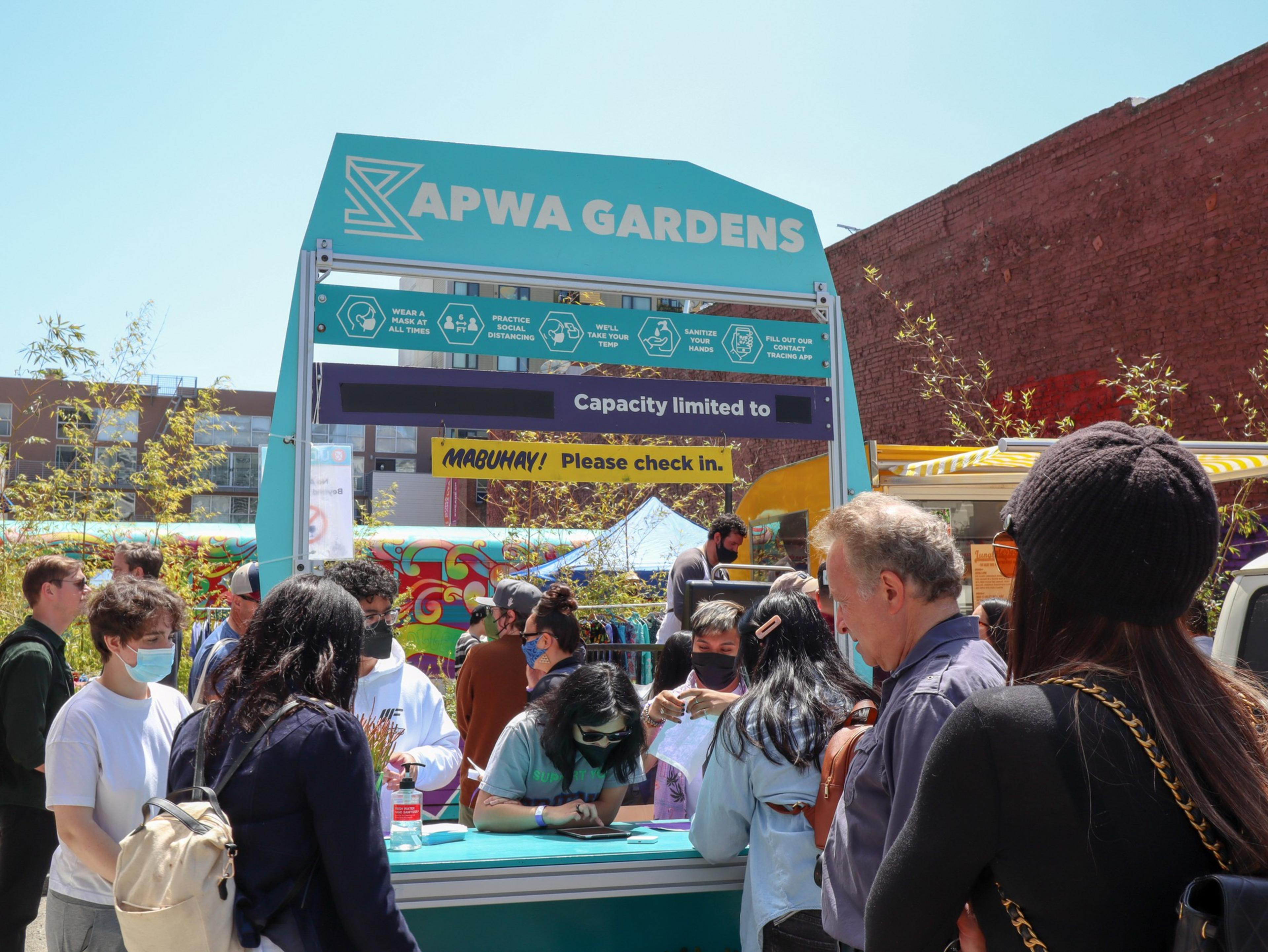 People wearing masks line up at a check-in booth under a sign reading "Sapwa Gardens" with COVID-19 safety reminders and a capacity limit note.