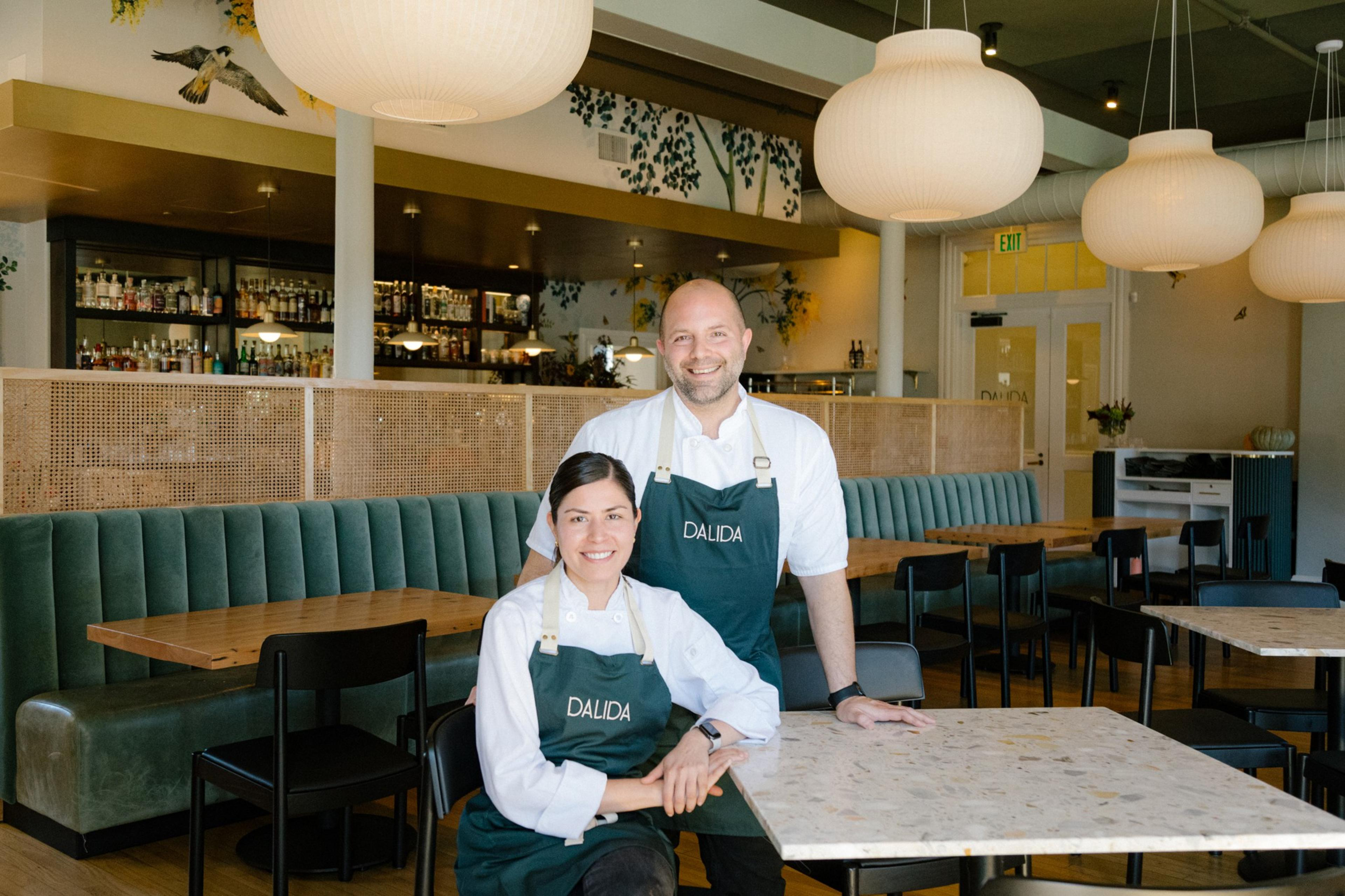 A smiling man and smiling woman, who are Executive chefs Laura and Sayat Ozyilmaz at Dalida, stand in the center of a modern restaurant that has large marble and wood tables spread throughout the dining space, circular lampshades on the ceiling and a bar i