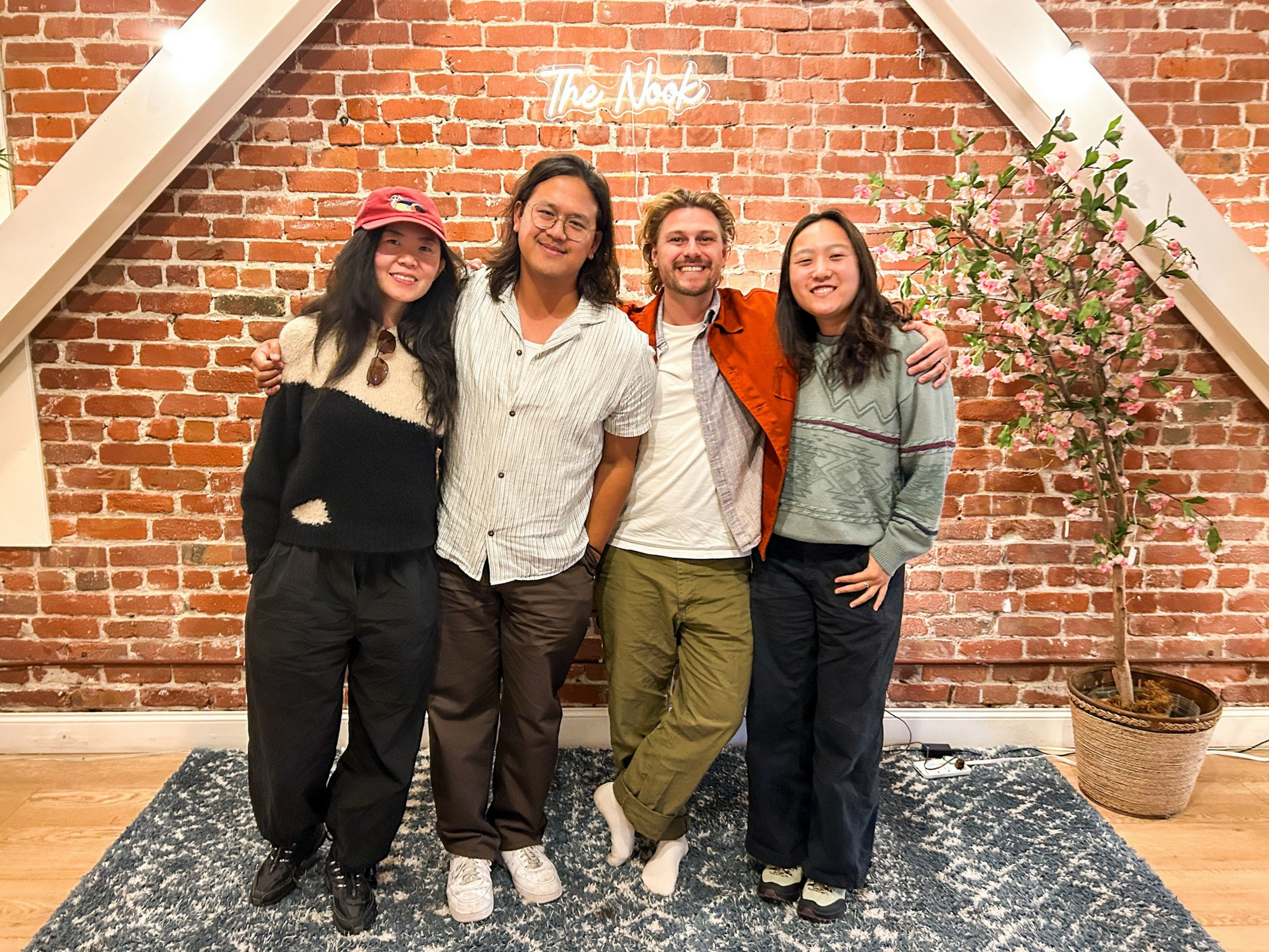 Four people stand arm in arm smiling in front of a brick wall with a neon sign reading “The Noob” and a potted flowering tree on the right.