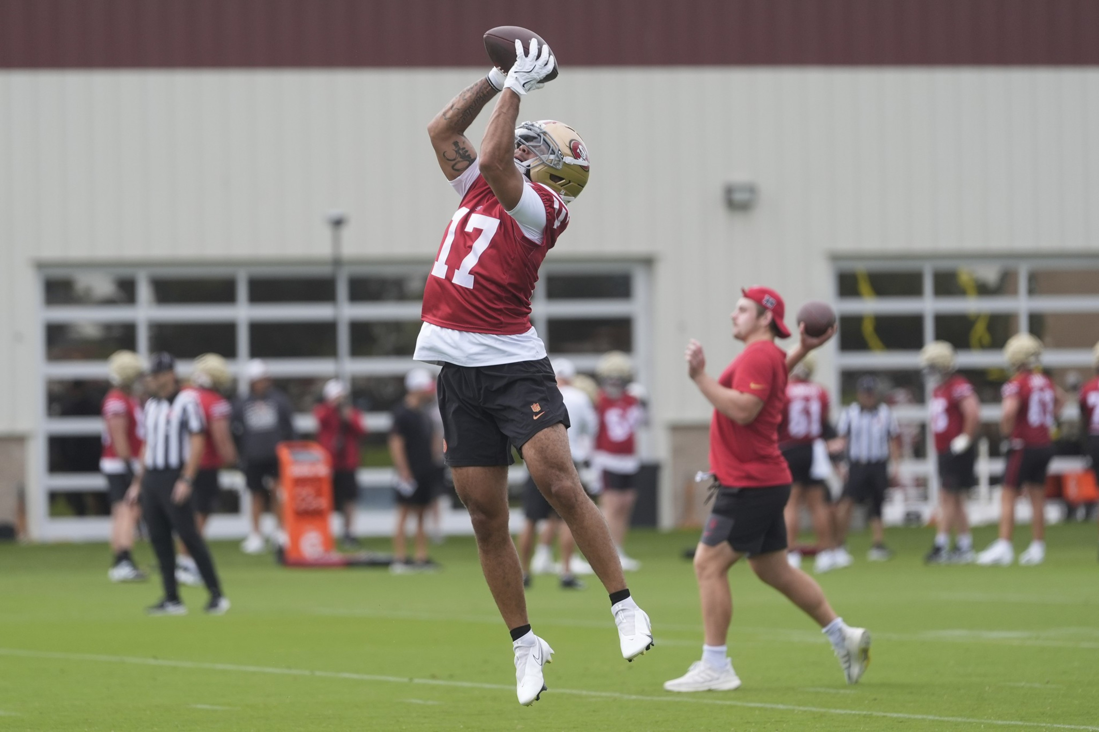 A football player in a red jersey leaps to catch a pass, while others practice in the background on a field.