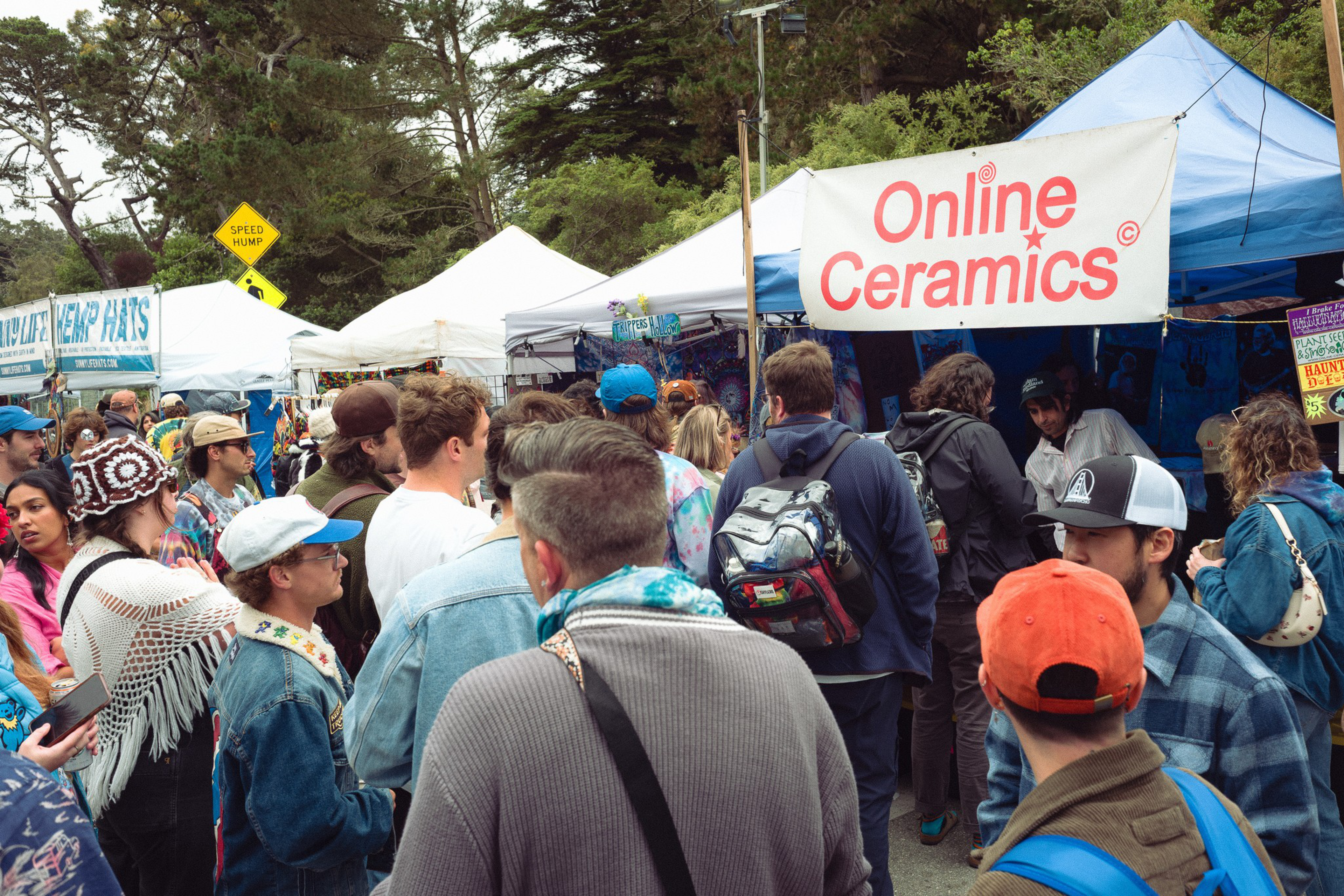 A crowded outdoor market features people browsing tents, including one labeled "Online Ceramics," with trees and a speed hump sign in the background.