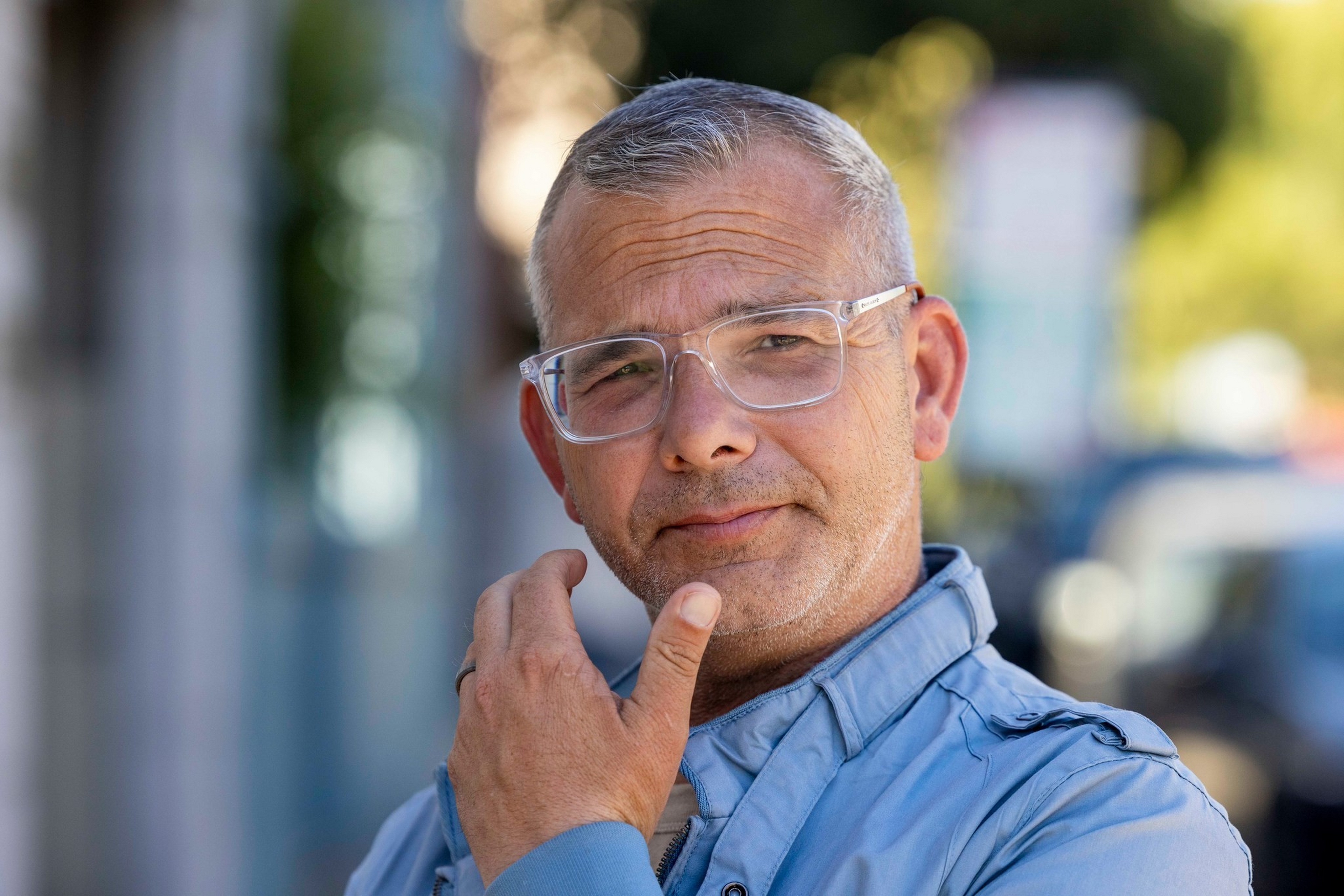 A middle-aged man with short gray hair and clear glasses touches his chin thoughtfully, wearing a blue jacket, with a blurred outdoor background.
