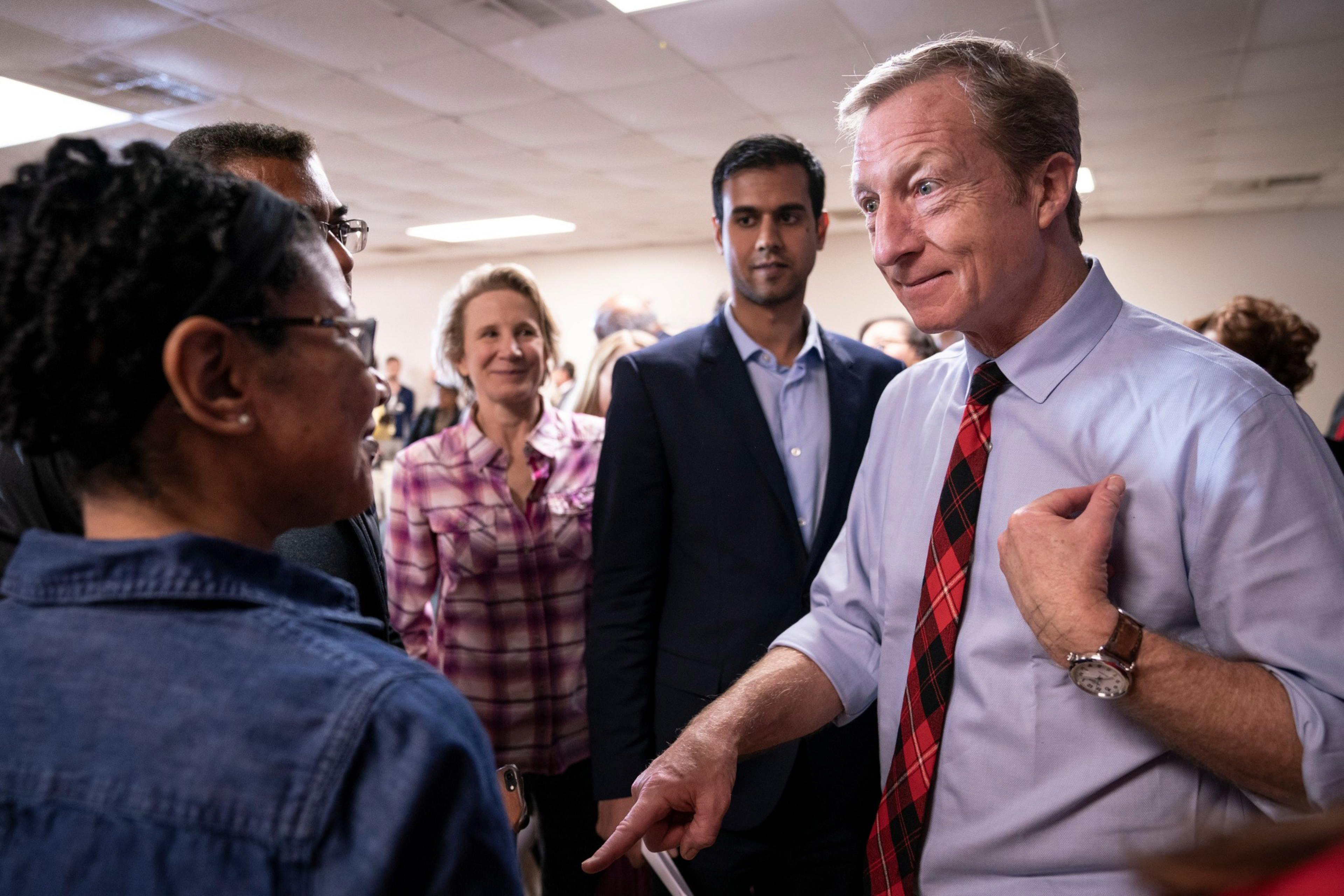 A man in a white shirt and red plaid tie speaks to a group of people, including a woman in glasses and a plaid shirt, in a room with a drop ceiling.