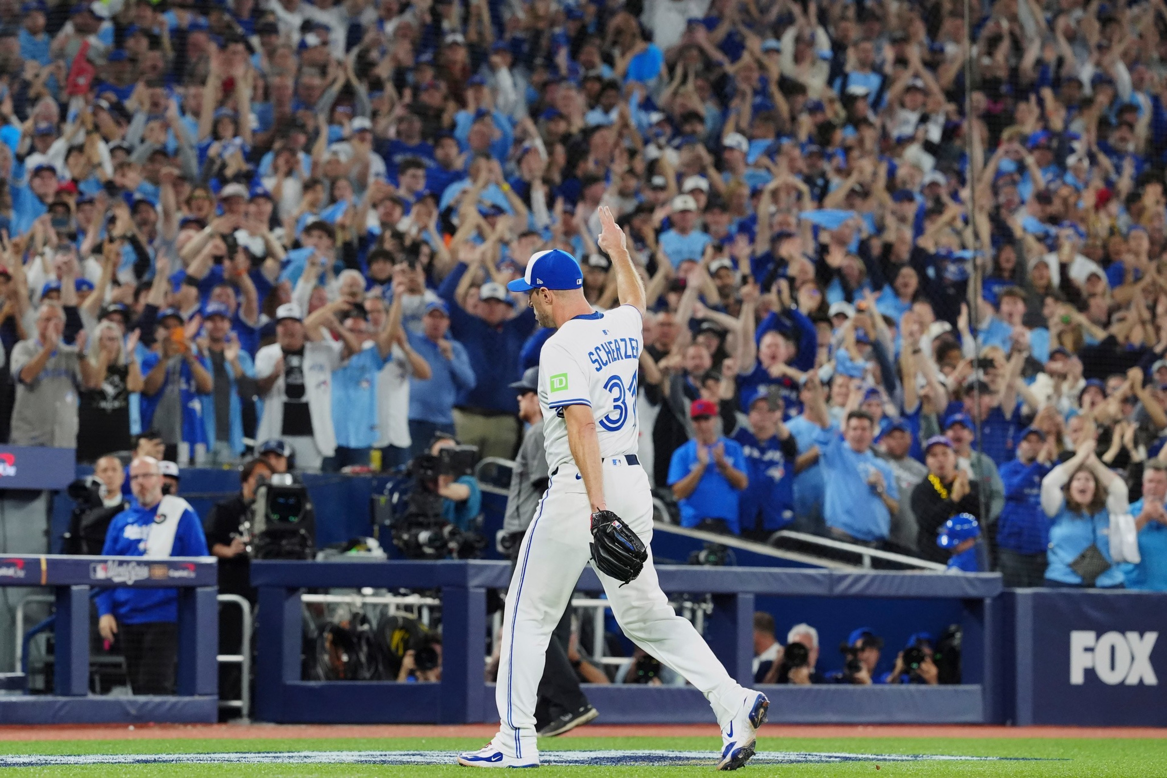 A baseball player wearing a white and blue uniform with “Scherzer 31" raises one hand while a crowd in blue cheers behind him.