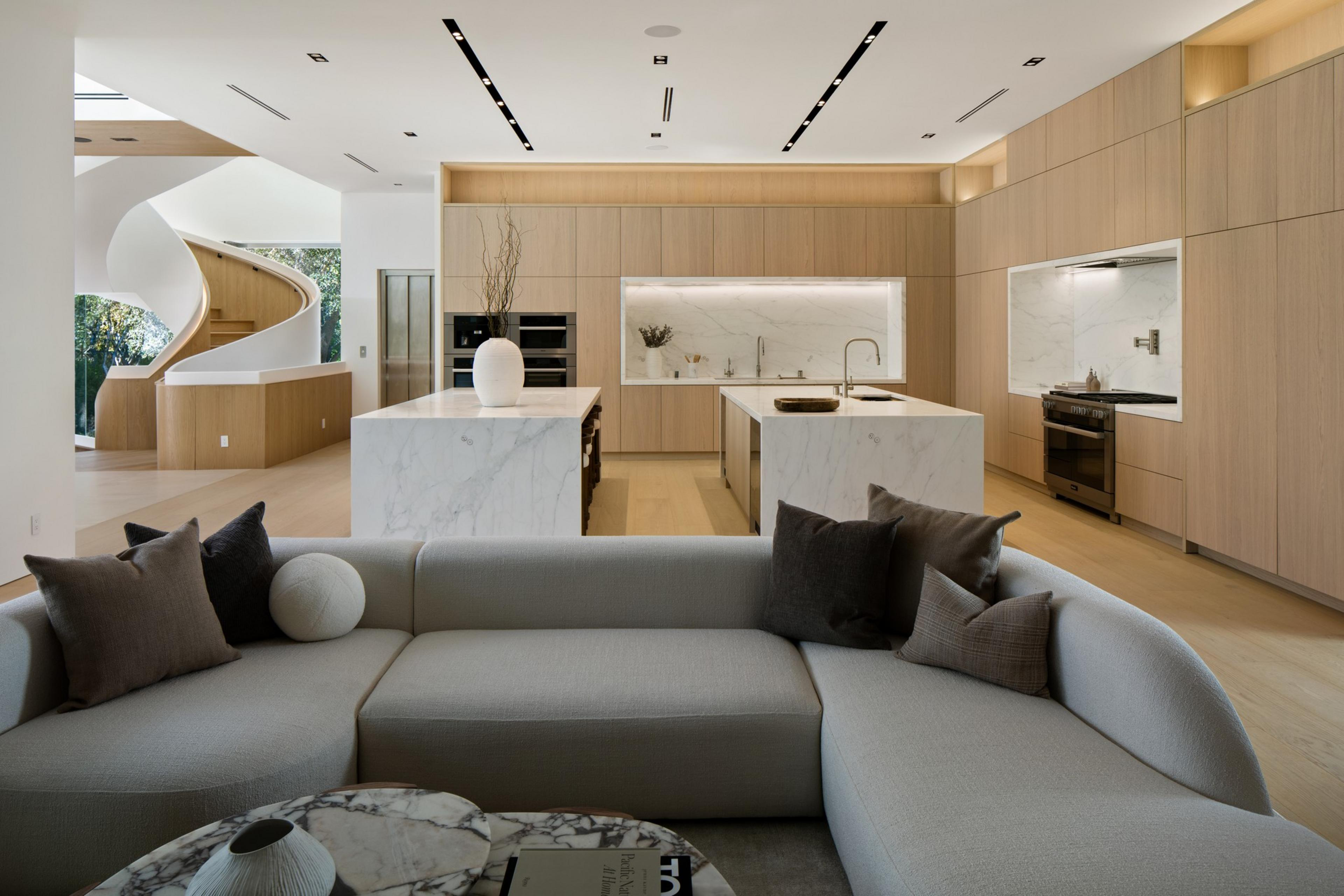 A modern kitchen and living room with light wood cabinets, marble countertops, a curved gray sofa, spiral staircase, and minimalist decor.