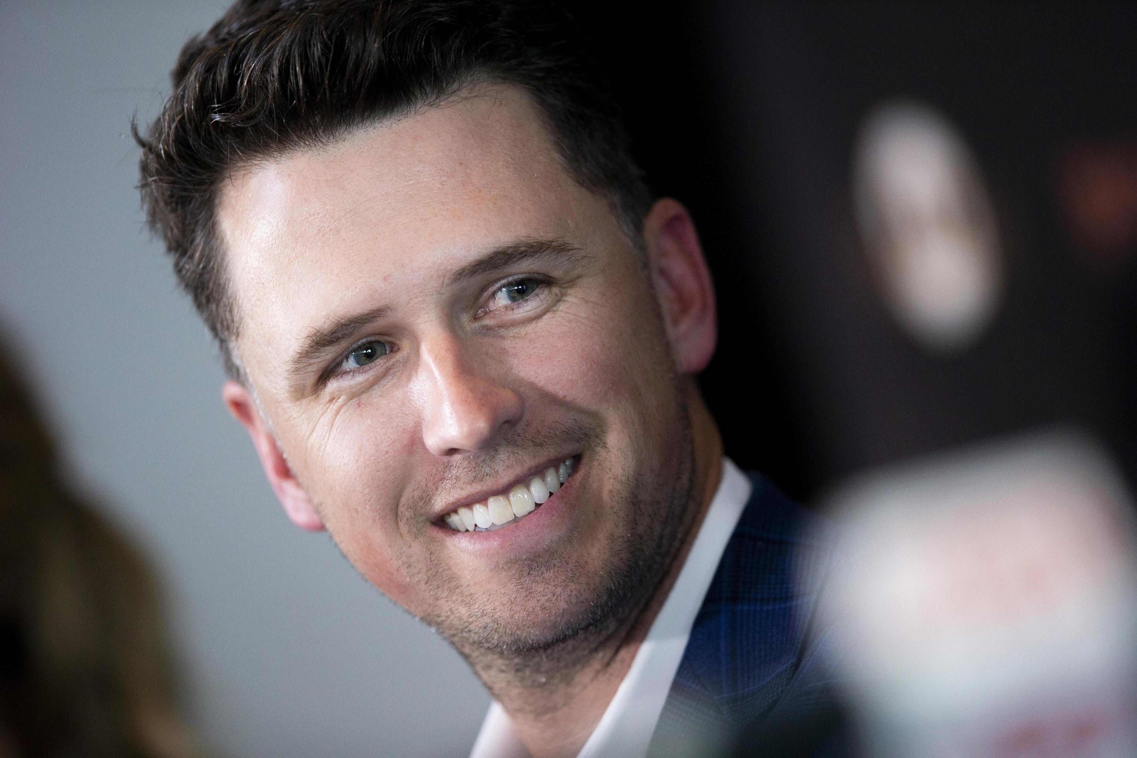 A man with short dark hair is smiling, looking to the side. He is wearing a suit, and the background is blurred with some indistinct objects.