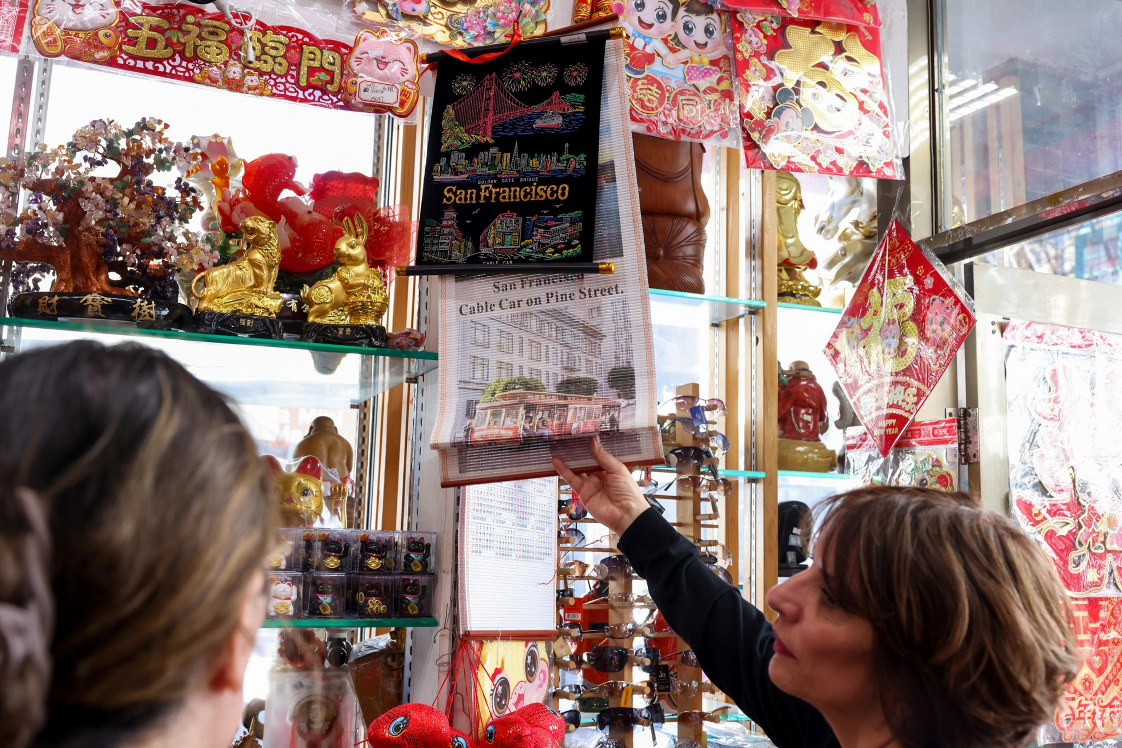 In a shop, a person holds a San Francisco tapestry featuring a cable car. Shelves display colorful ornaments, golden animal figurines, and Chinese decorations.