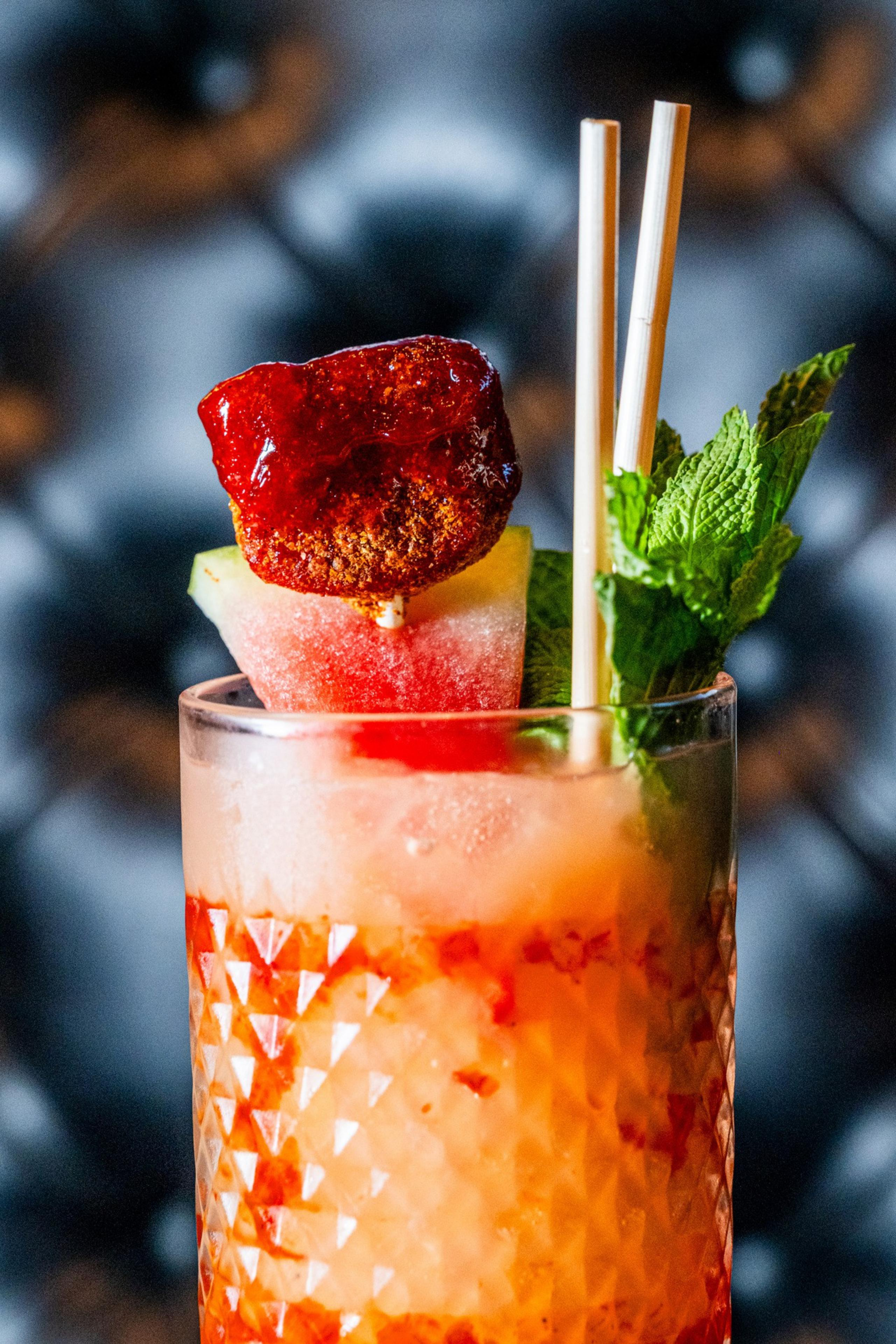 A textured glass contains a vibrant drink garnished with a red candy, watermelon slice, mint leaves, and two straws, set against a blurred background.