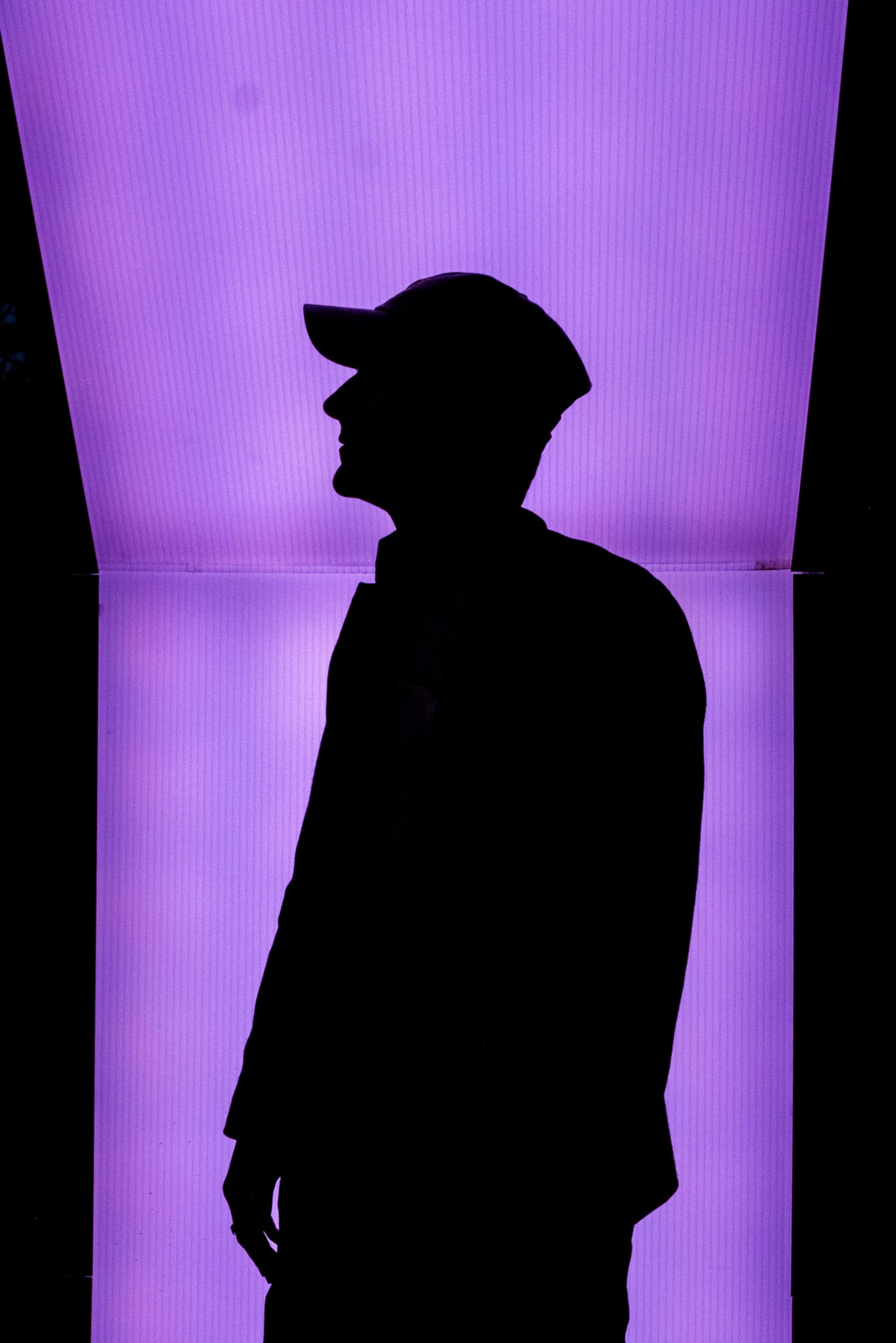A silhouette of a person wearing a cap is seen against a bright purple illuminated background.
