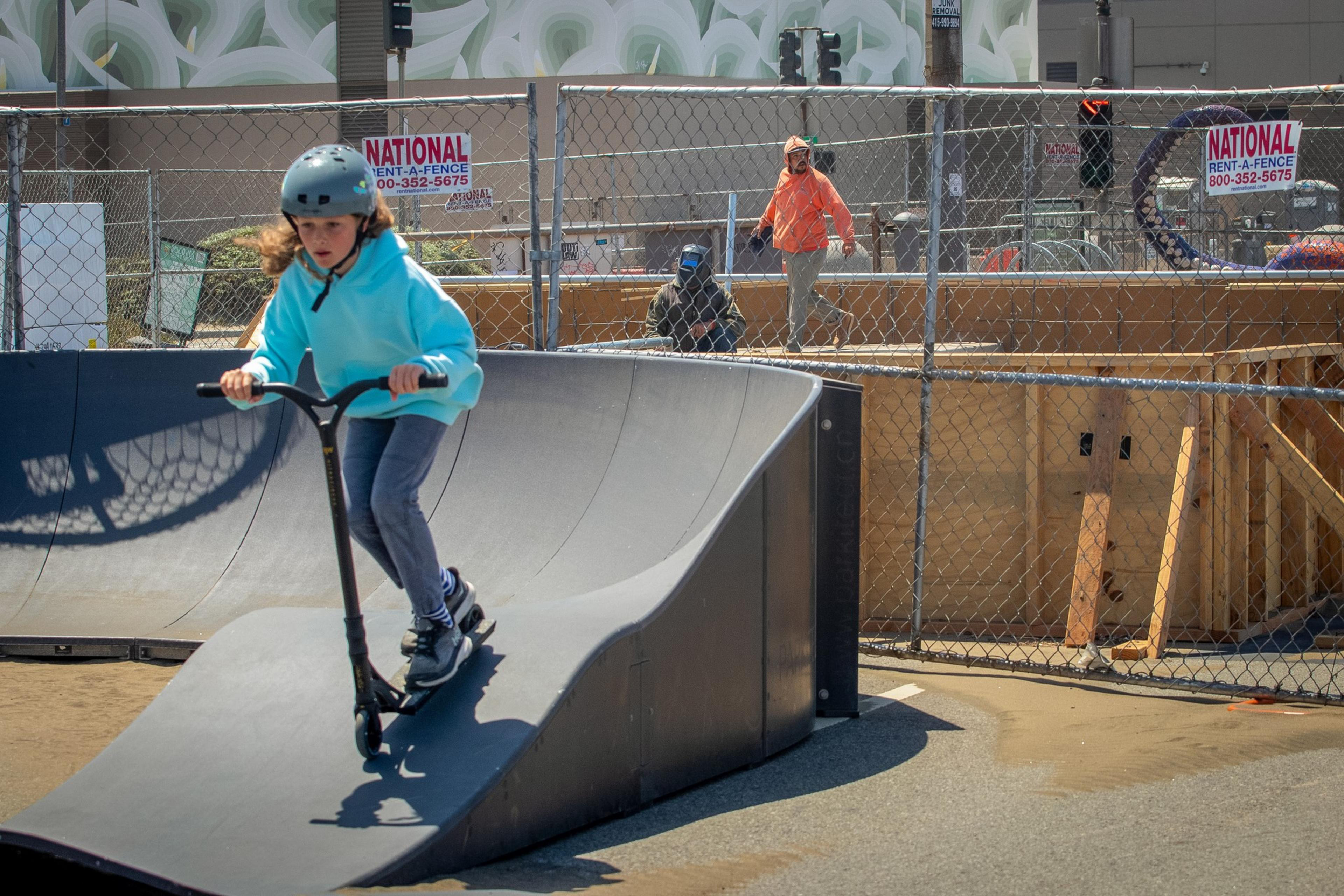 A child wearing a helmet and light blue hoodie rides a scooter down a ramp in a skate park. A fence with signs and workers in safety vests are in the background.