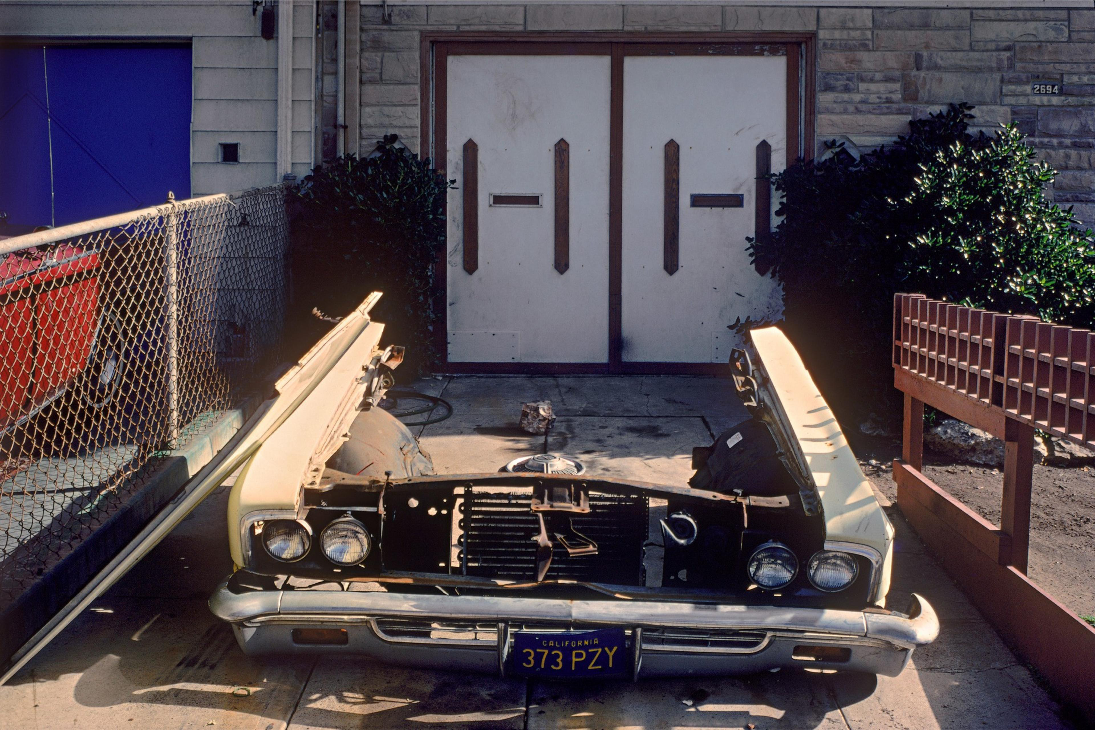 A dismantled car front is placed in a driveway, framed by a chain-link fence on the left and a wooden fence on the right, in front of double garage doors.