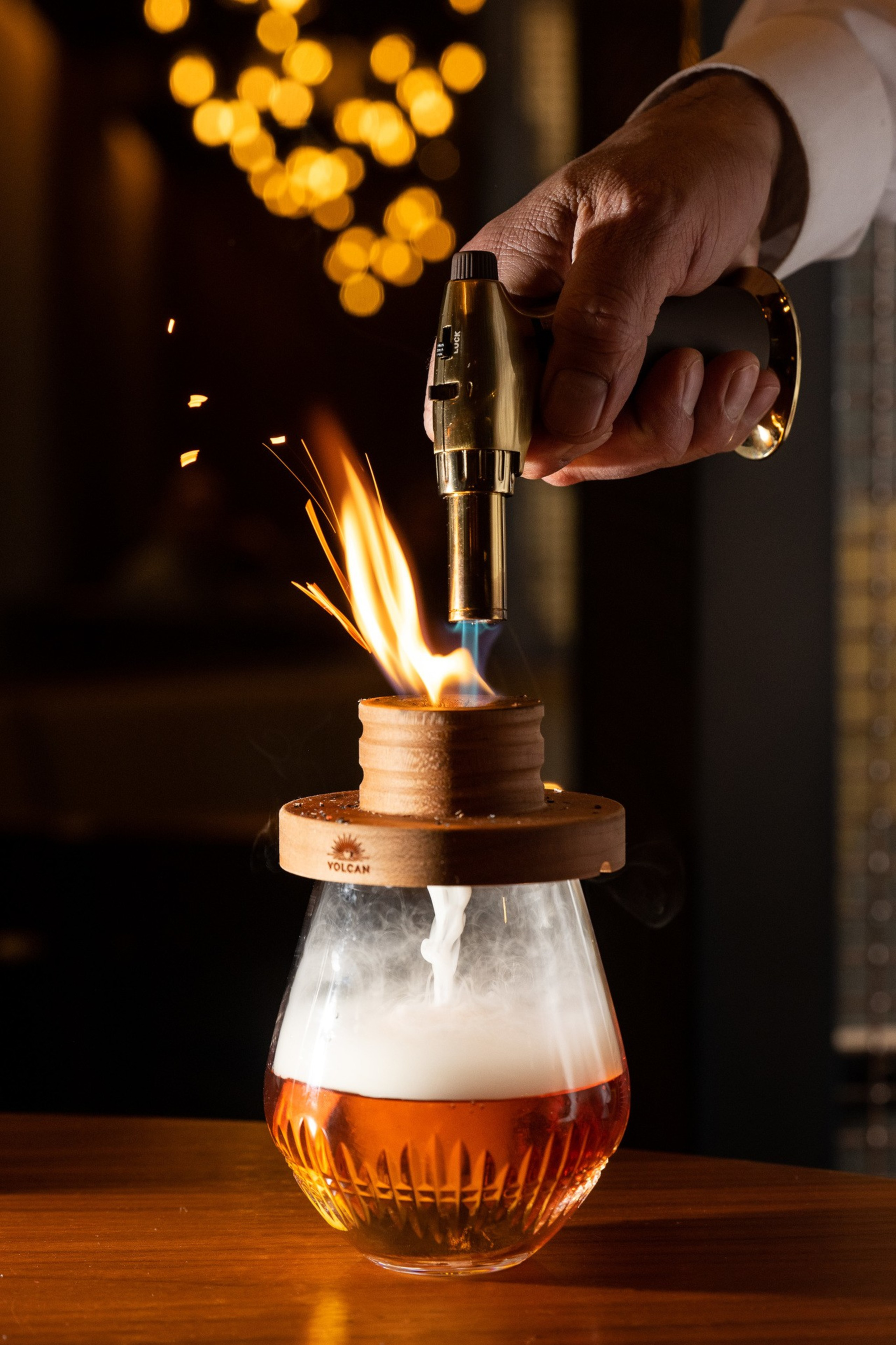 A hand holds a torch flame over a smoking drink in a glass with a wooden lid, creating sparks and smoke rising from the cup.