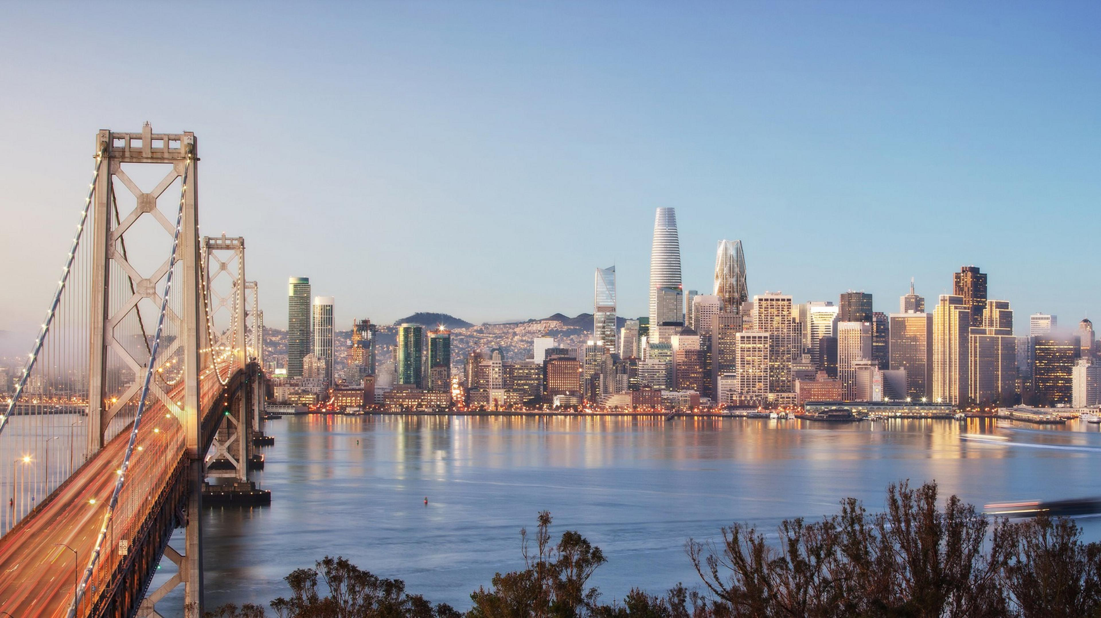 A bridge leads to a city skyline at dusk with illuminated buildings reflecting on the water.