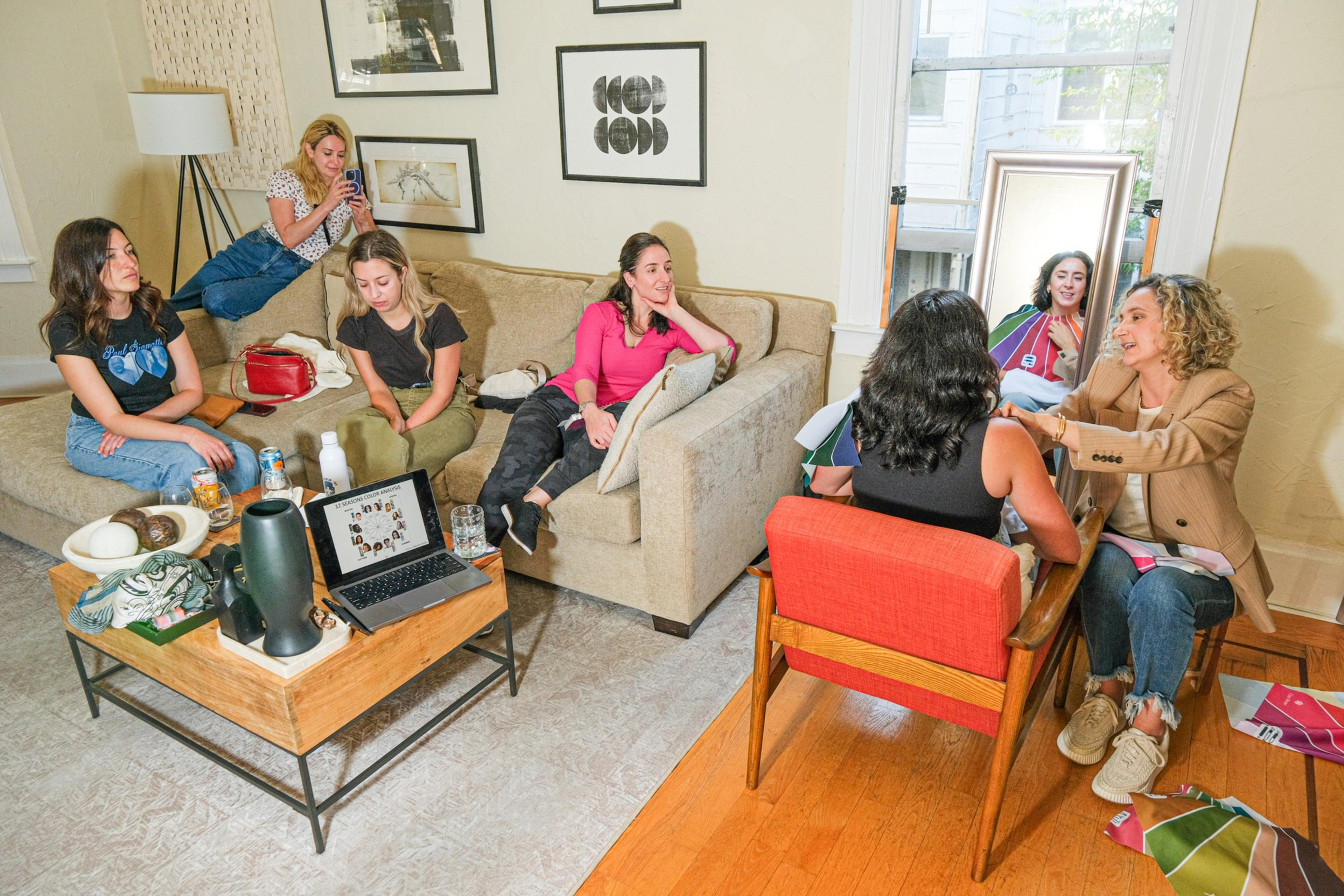 Six women are in a cozy living room. Some are chatting, while one takes photos and another helps another woman with colorful fabrics by a large mirror.