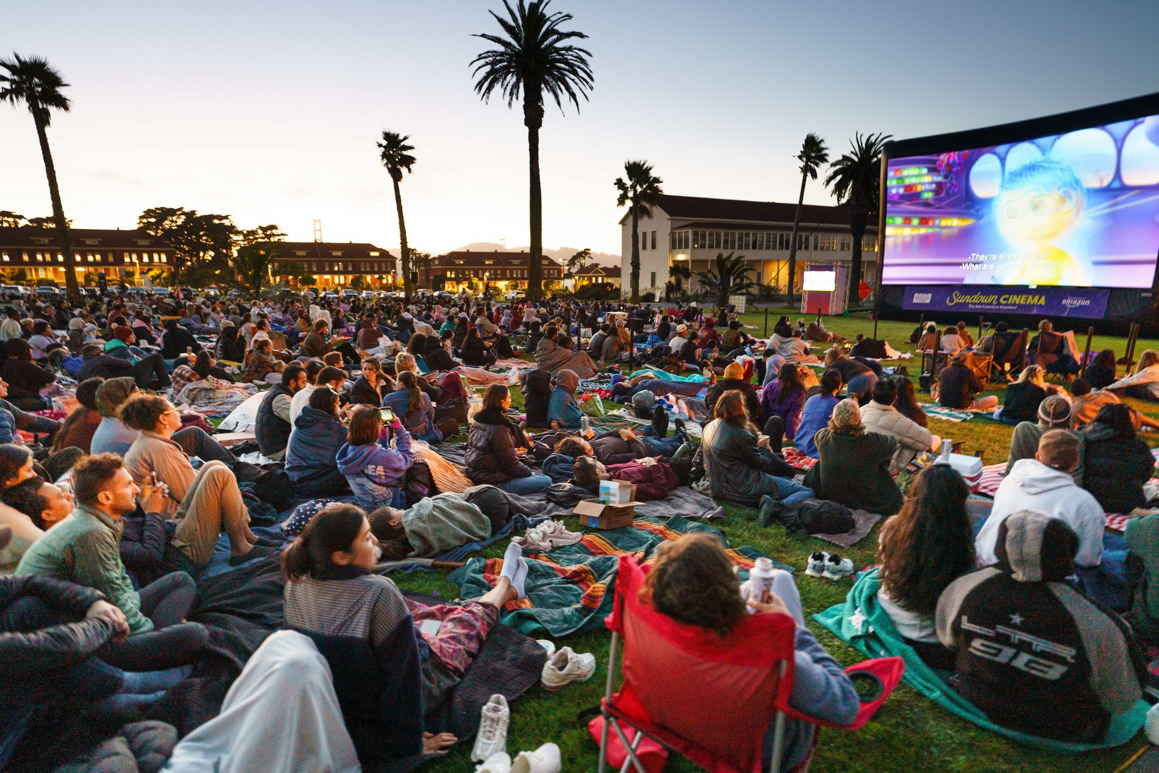 A large crowd sits on blankets and chairs on grass, watching an animated movie at an outdoor cinema during sunset, with palm trees and buildings in the backgrou