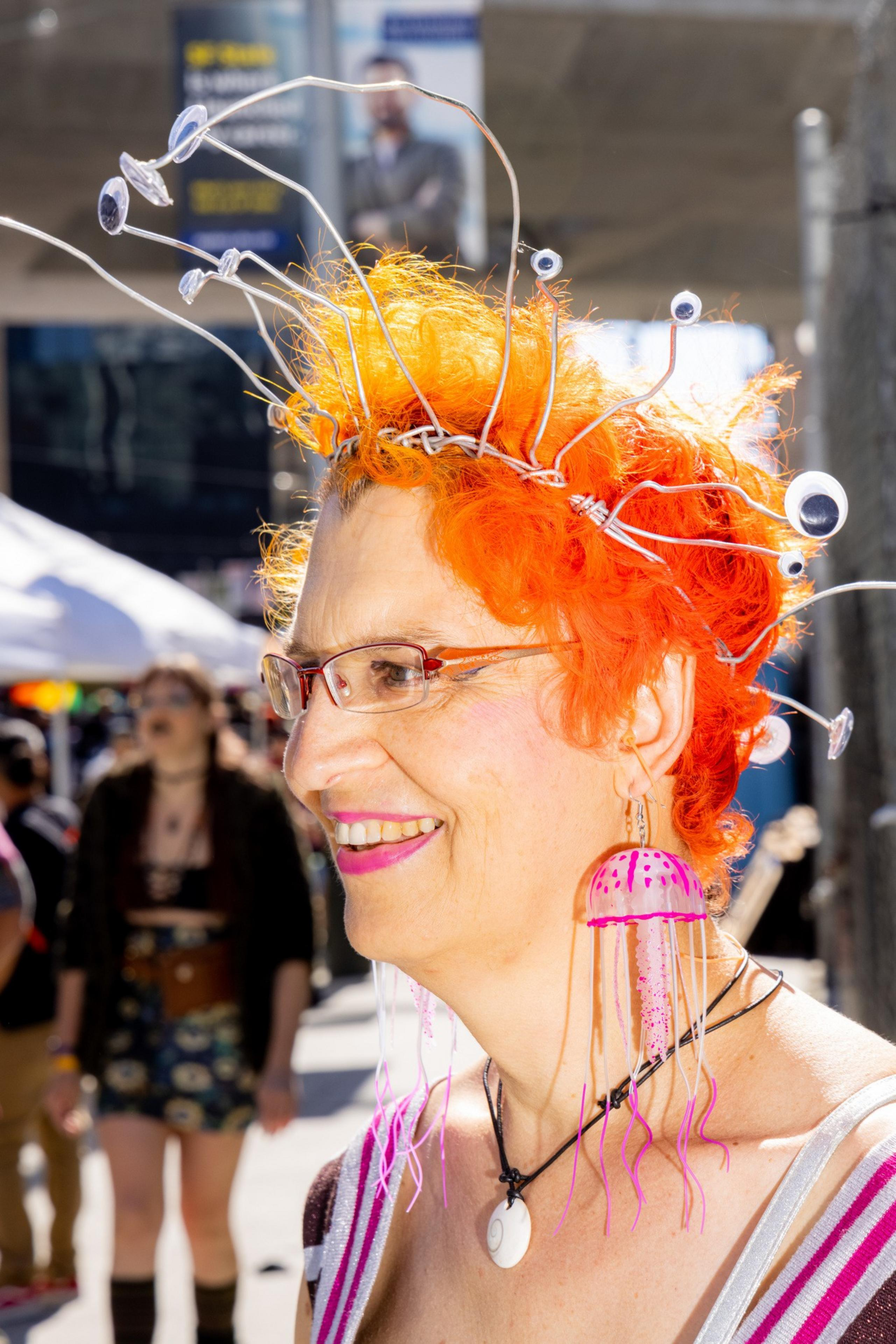 A person with bright orange hair, adorned with a whimsical wire headpiece and googly eyes, smiles at an outdoor event. They wear large, colorful jellyfish earrings and glasses.