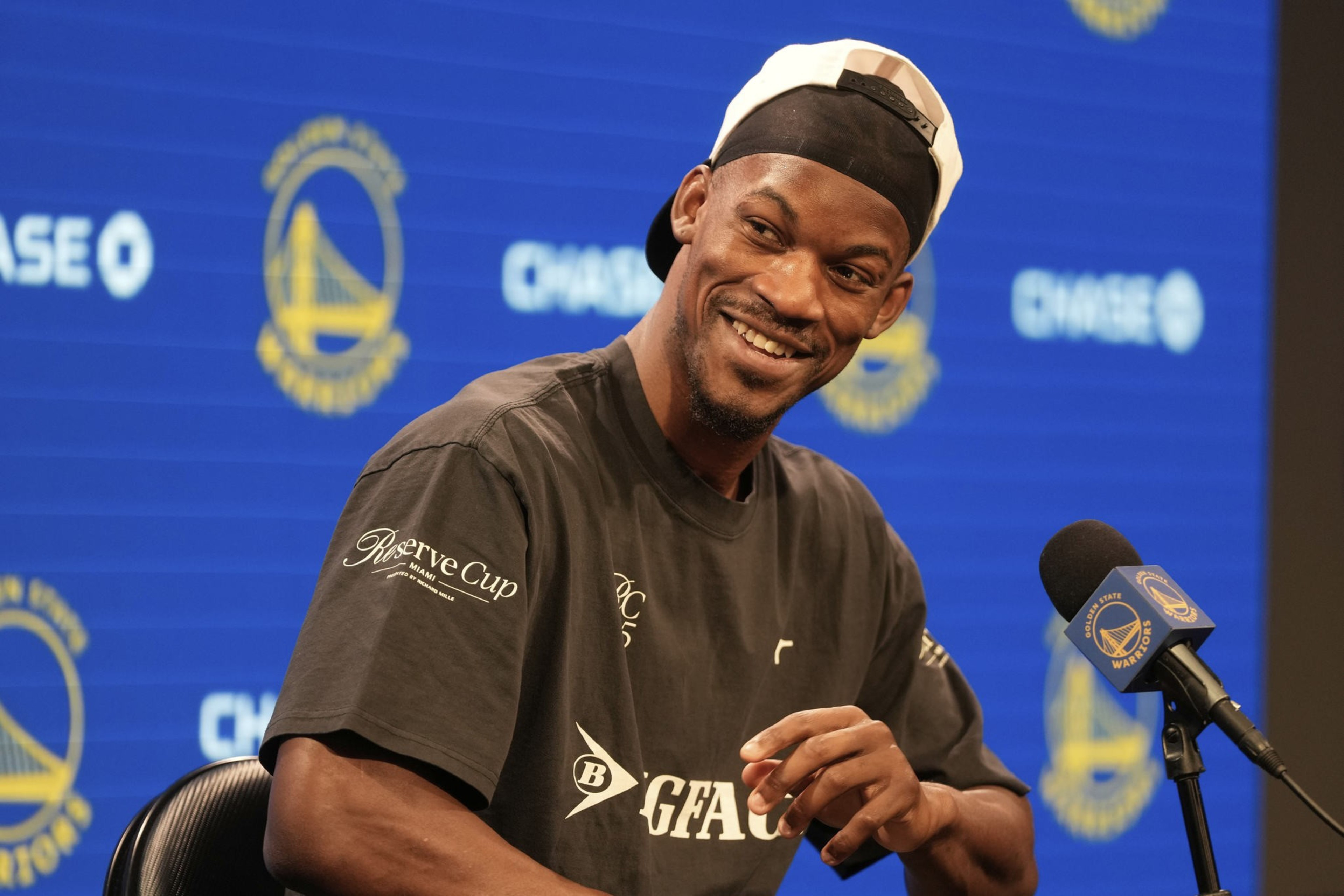A man in a black shirt and backwards cap smiles at a press event with a Golden State Warriors backdrop. A microphone is in front of him.