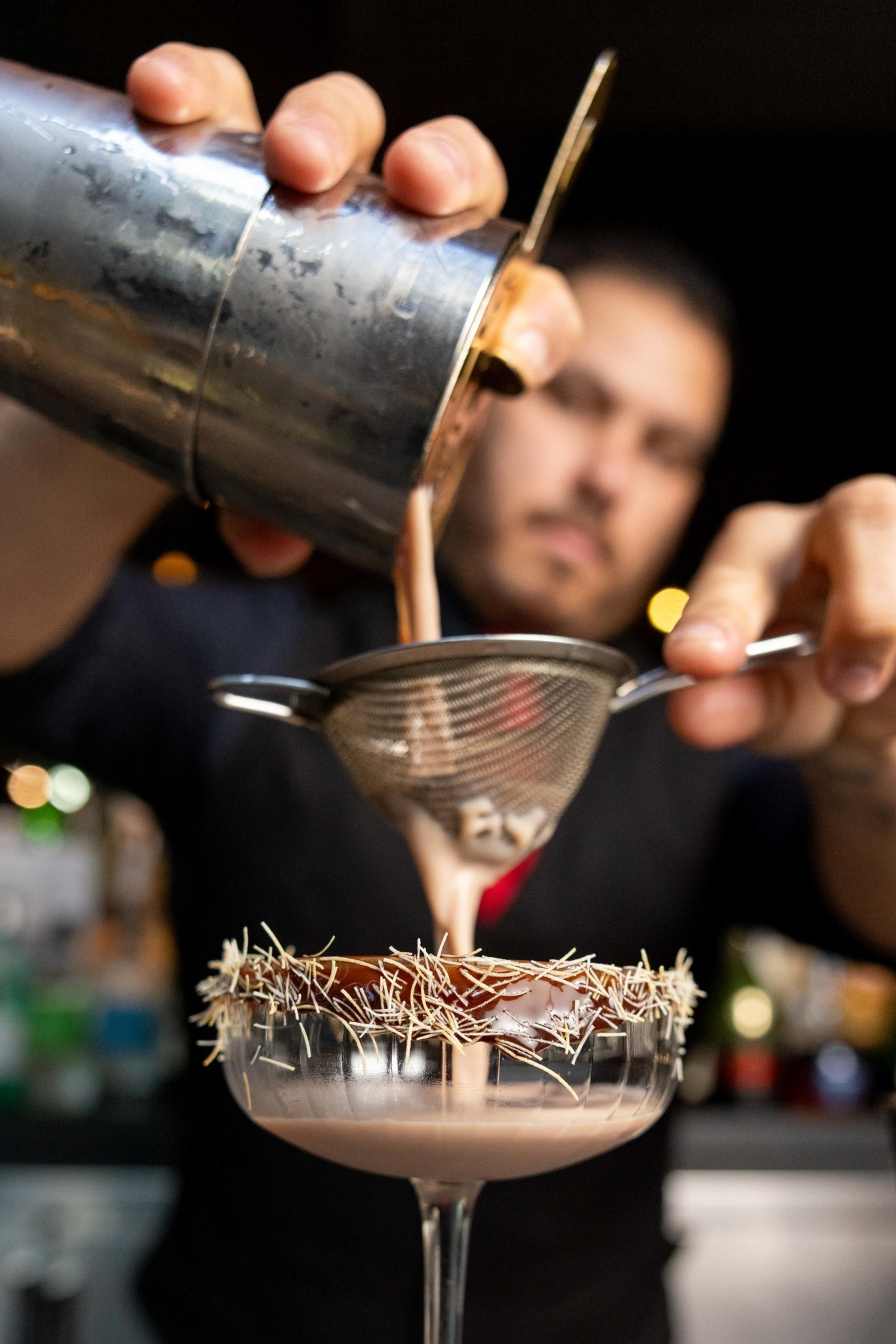 A bartender is pouring a creamy cocktail through a strainer into a glass. The glass rim is decorated with fine chocolate shavings.