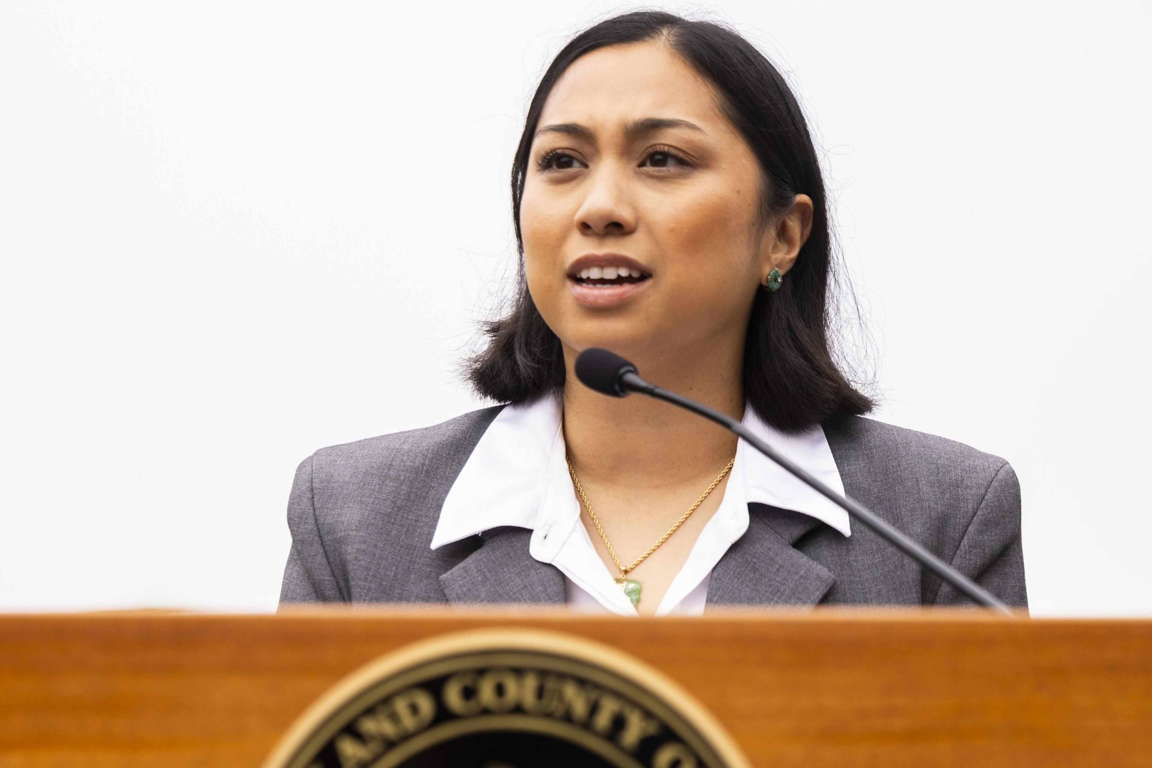A woman wearing a gray blazer and white shirt speaks at a podium with a microphone, appearing focused and engaged.