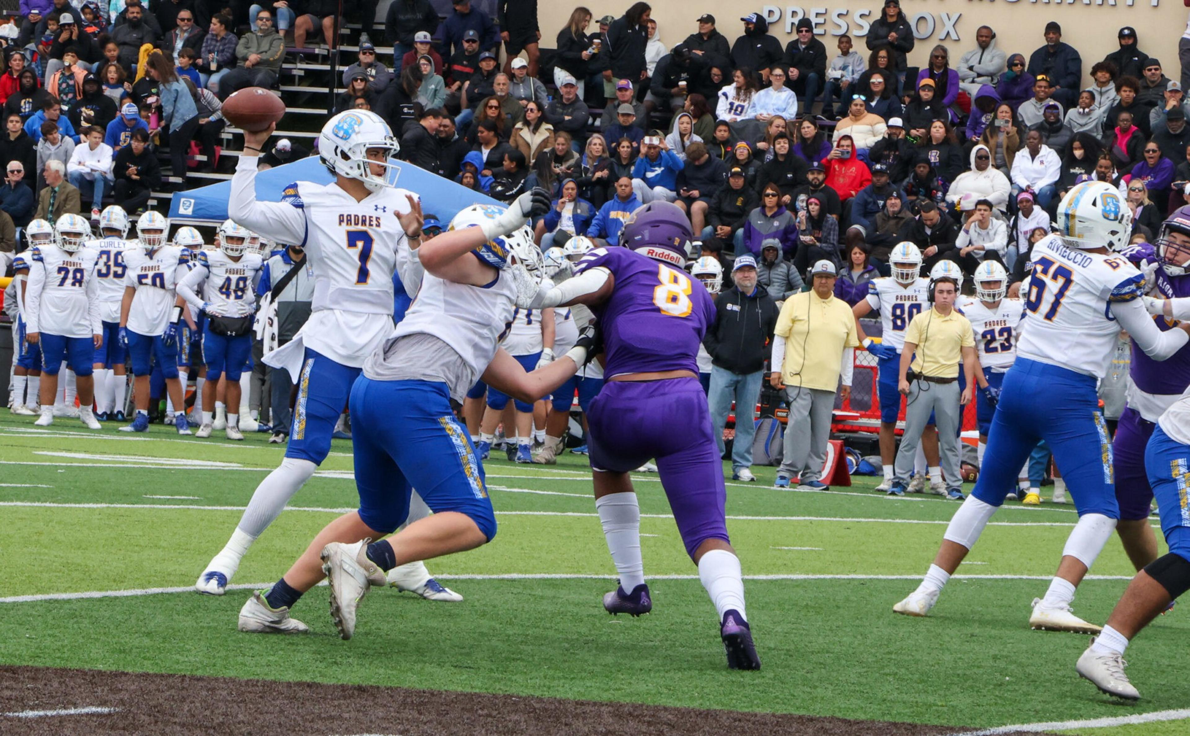 A football quarterback in white prepares to throw while players in white and blue block a player in purple on a crowded field with spectators in the background.