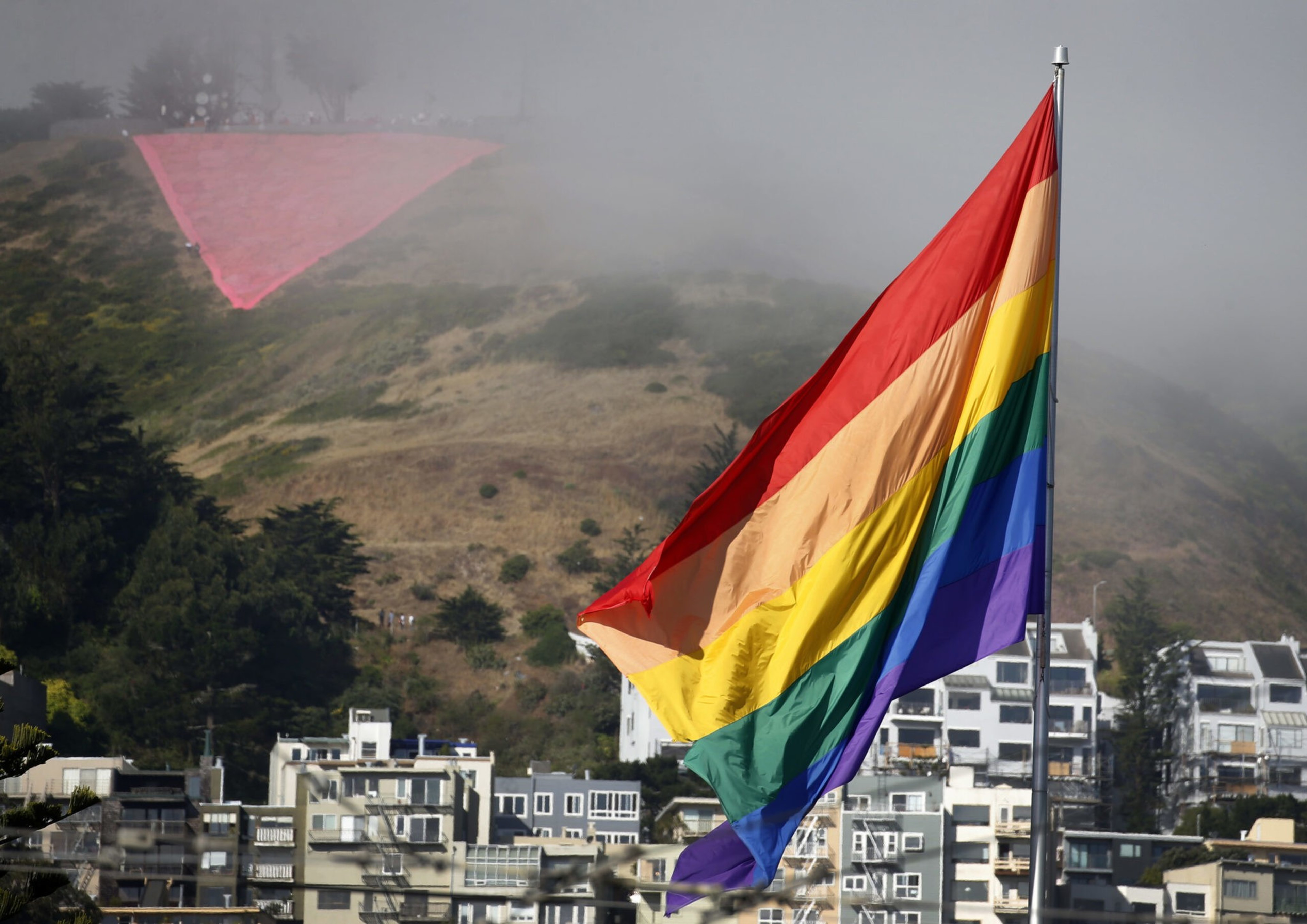 A large rainbow Pride flag flies with a foggy hillside and a red triangle painted on the hill above city buildings in the background.
