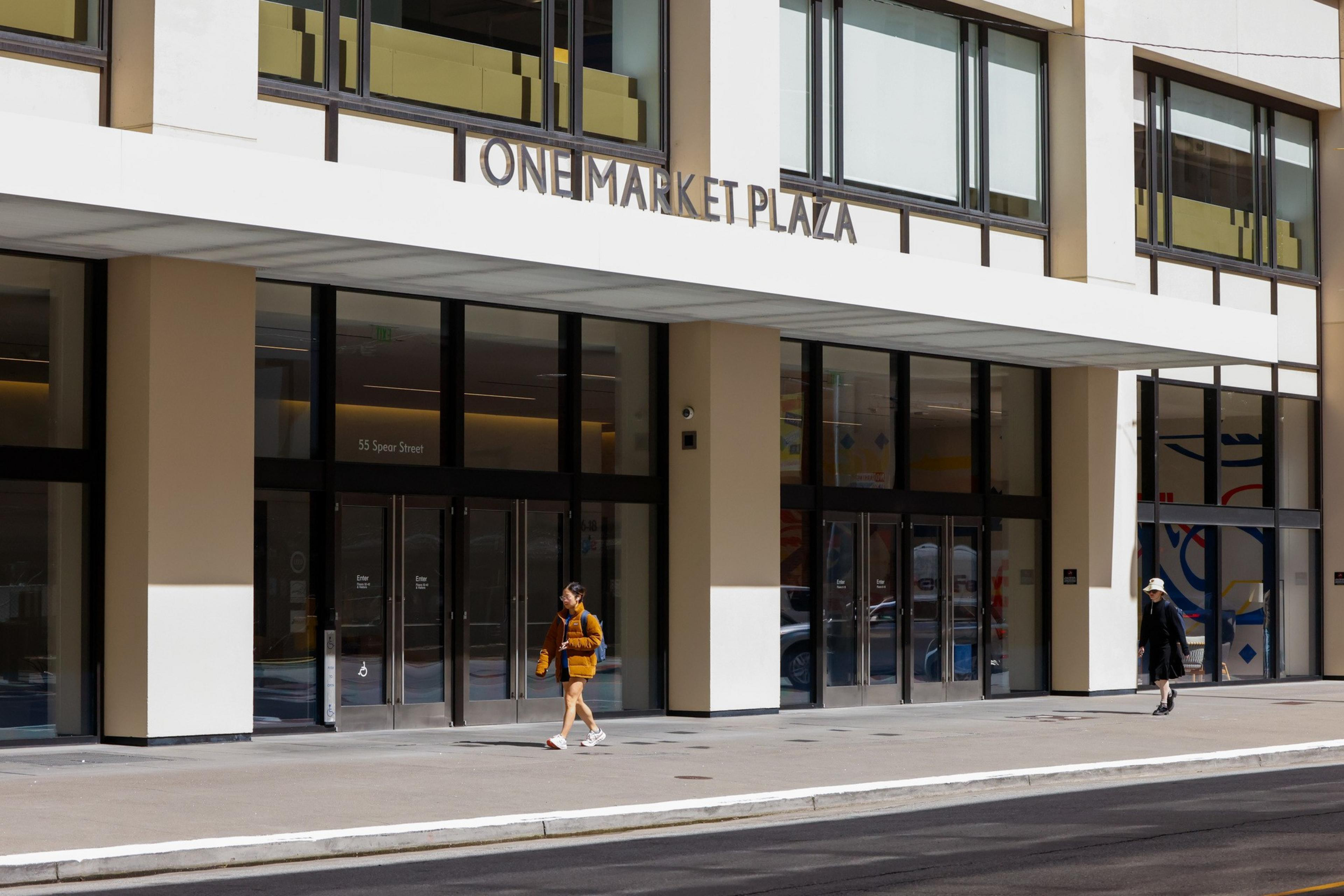 The image shows the entrance to One Market Plaza with two people walking by, one in a brown jacket and another in a hat. The building has large glass doors.