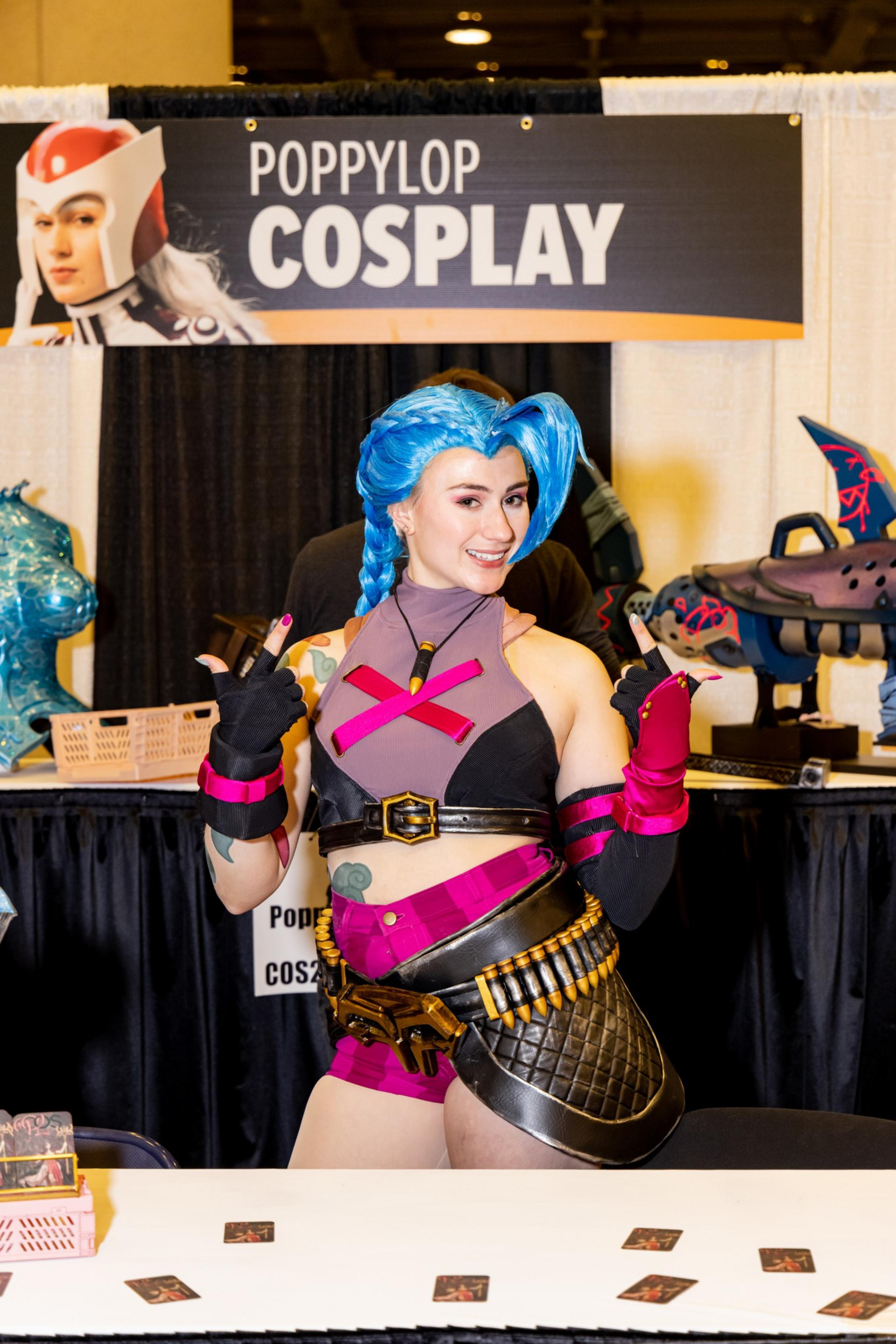 A person in a vibrant cosplay costume with blue hair and a punk-inspired outfit poses confidently in front of a booth sign reading &quot;Poppylop Cosplay.&quot;