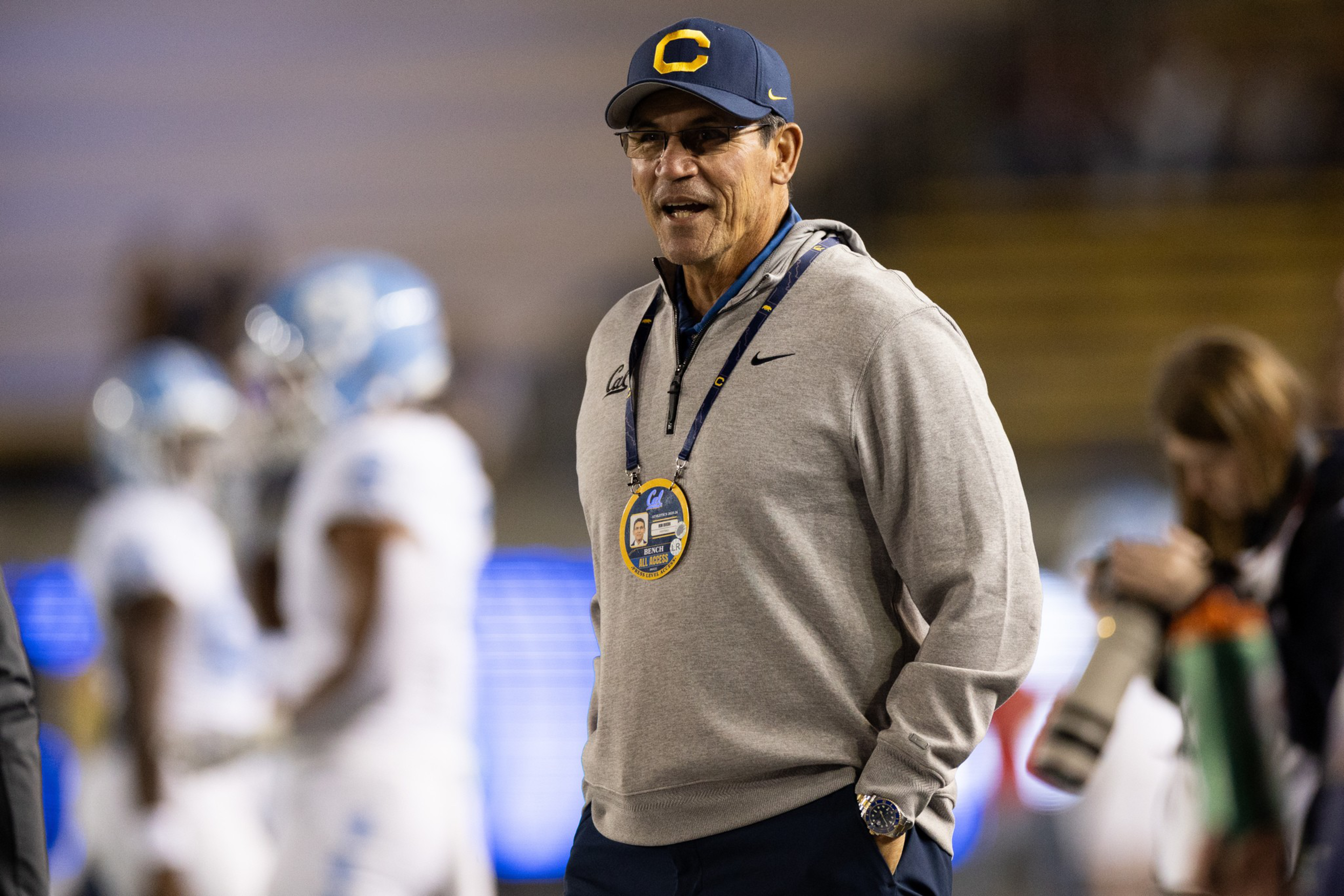 A man wearing a gray hoodie and navy hat with a “C” stands on a sports field, smiling with his hands in his pockets and a large ID badge around his neck.
