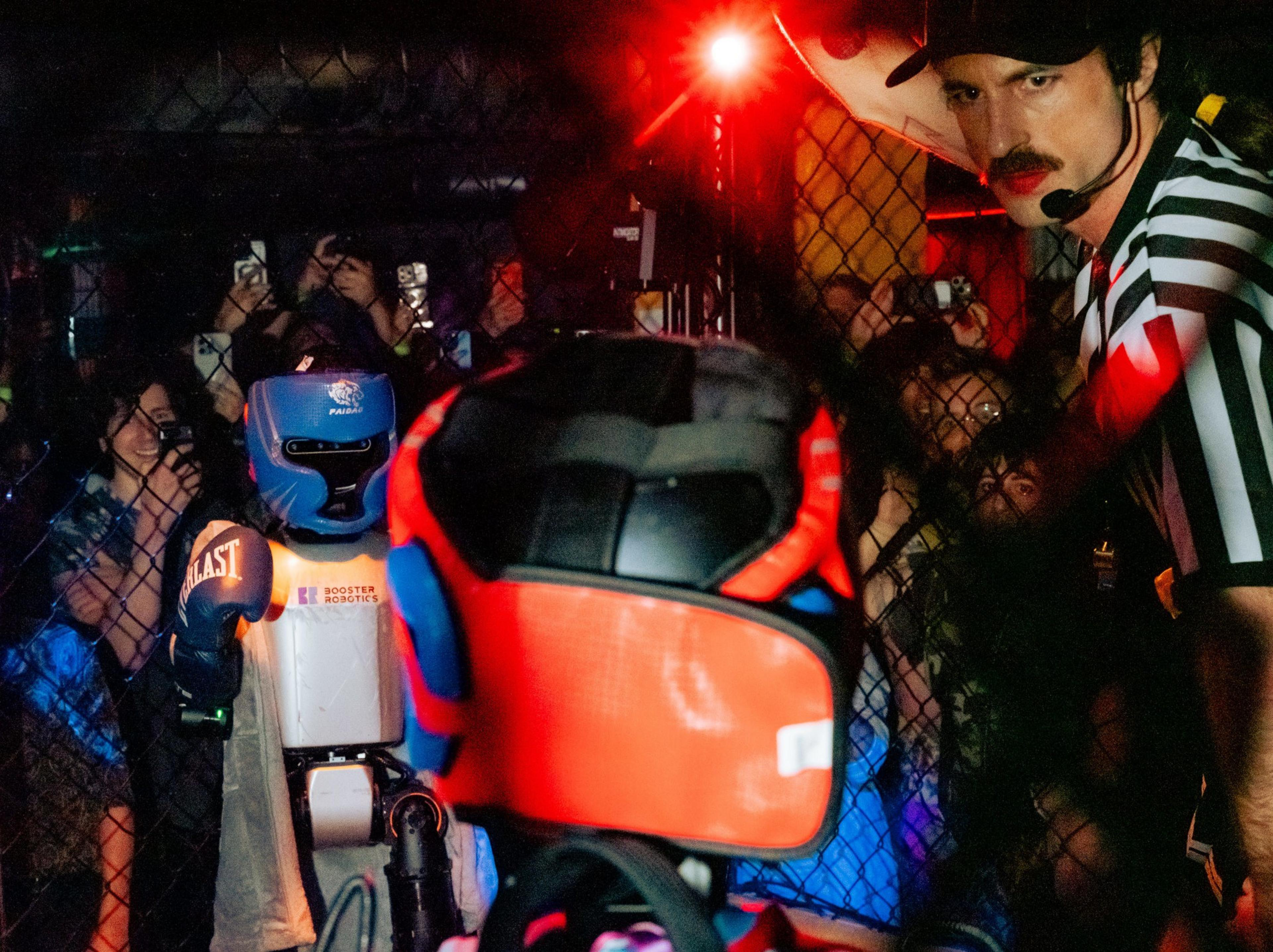 A robot wearing blue headgear and boxing gloves faces another robot with orange headgear inside a caged ring while a referee watches closely.