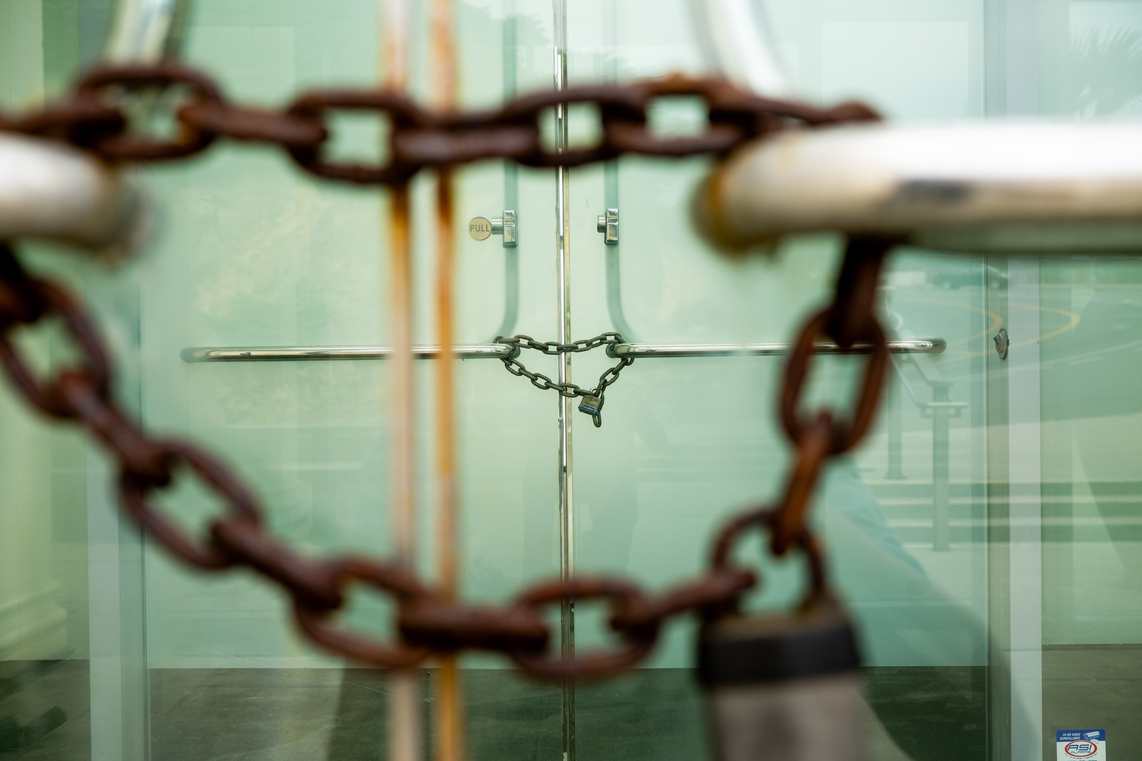 A glass door is secured with a chain and padlock, with an additional rusty chain and padlock visible in the foreground.