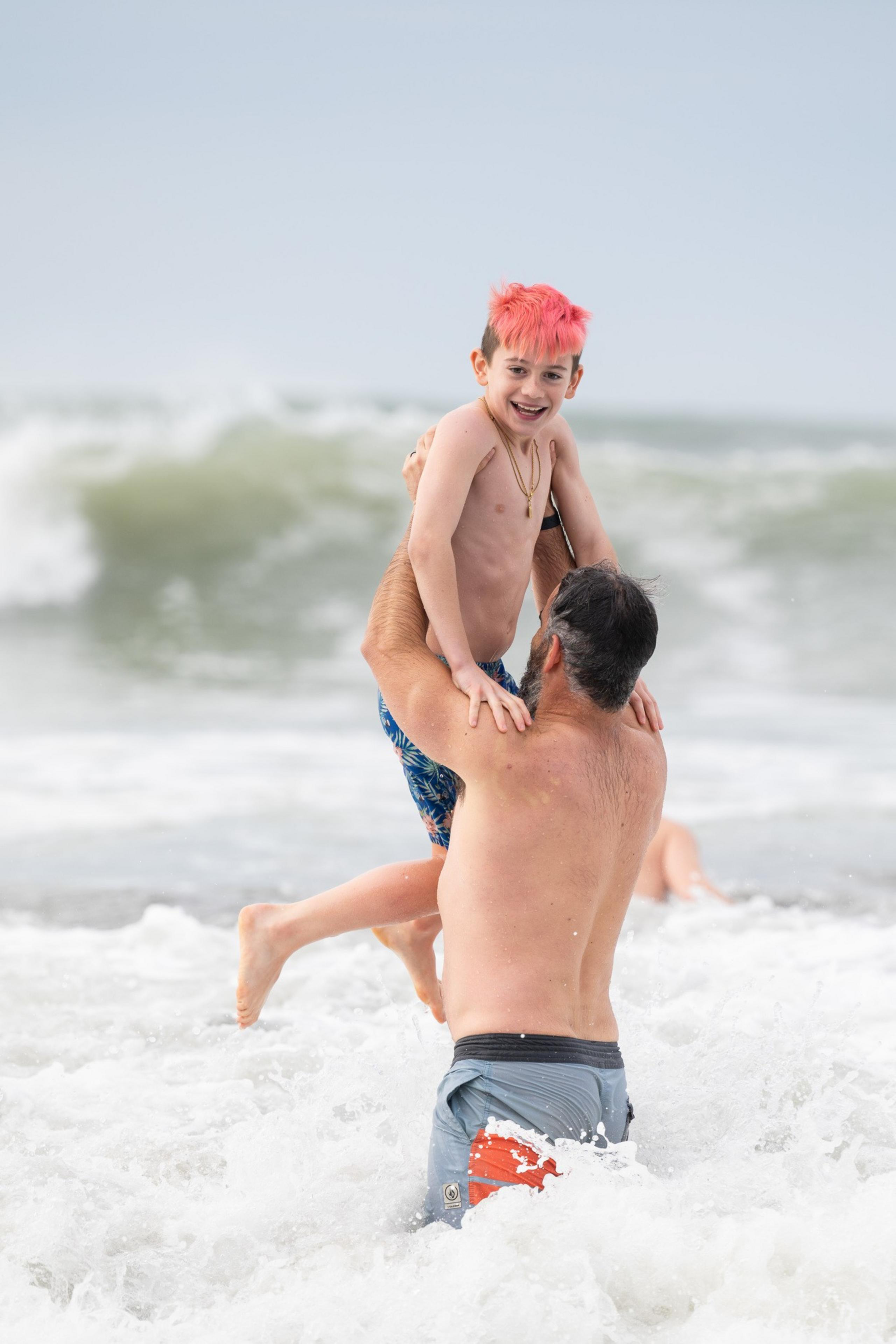 A man in swim trunks holds a smiling boy with bright pink hair above ocean waves, both enjoying a playful moment at the beach.