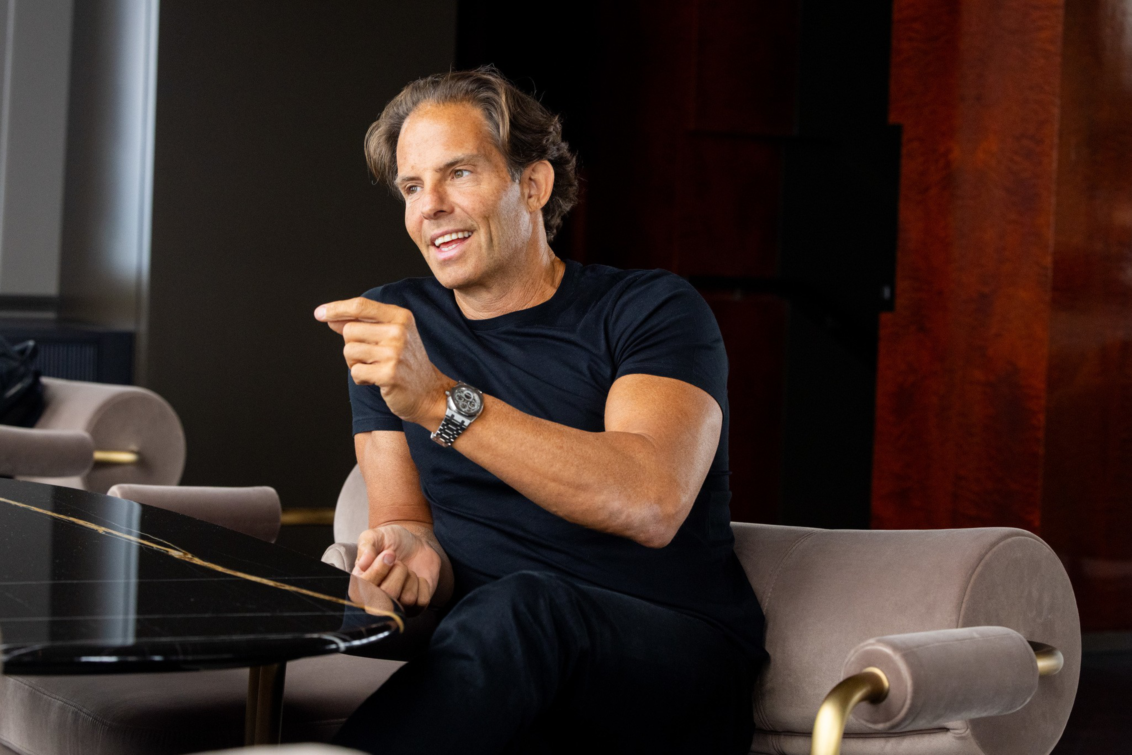 A man in a black shirt sits on a beige chair, gesturing with his right hand while talking, near a black table with gold veins.