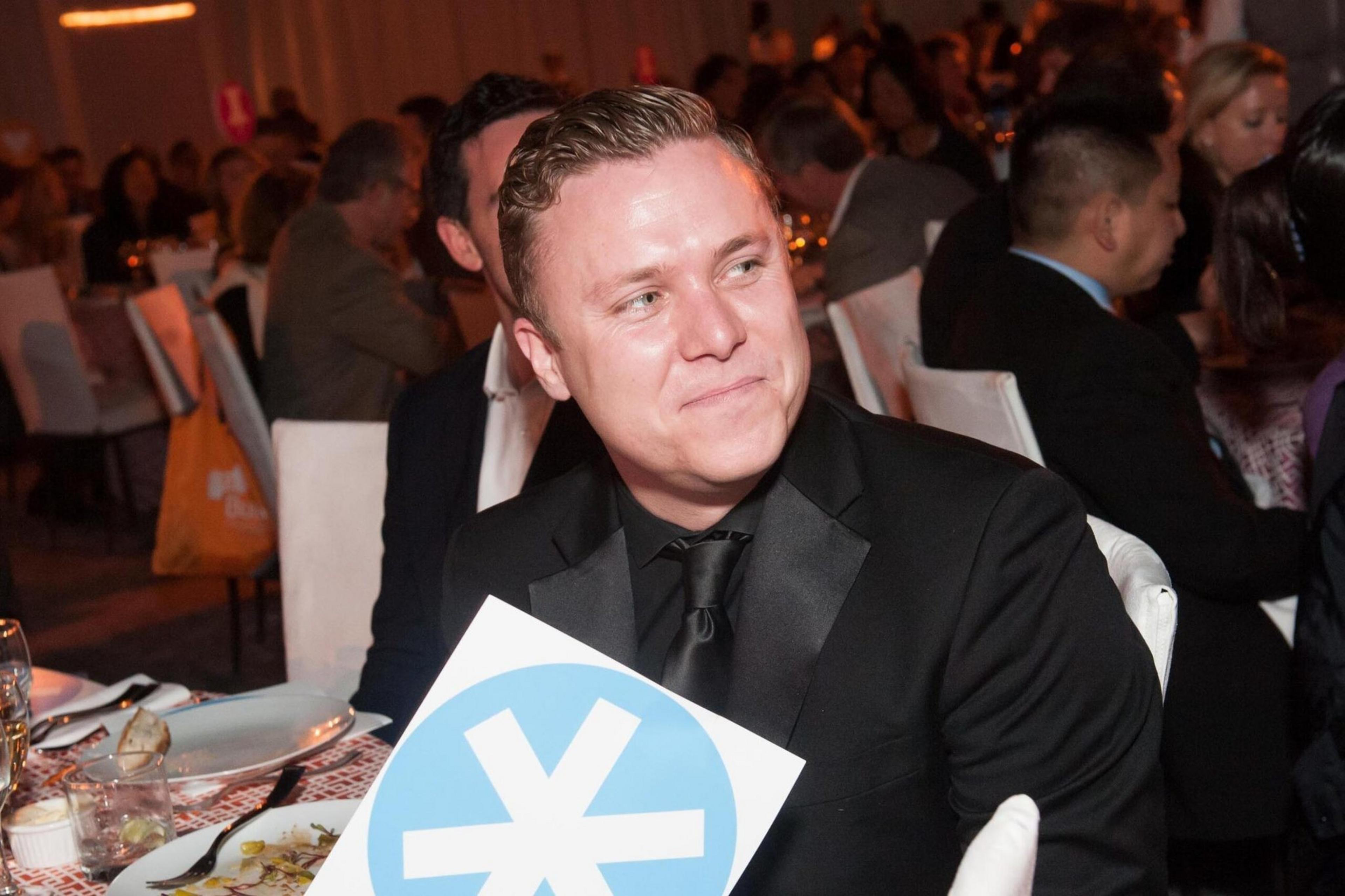 A man in a black suit and tie sits at a formal dinner, holding a sign with a blue circle and white asterisk-like symbol, smiling and looking to the side.