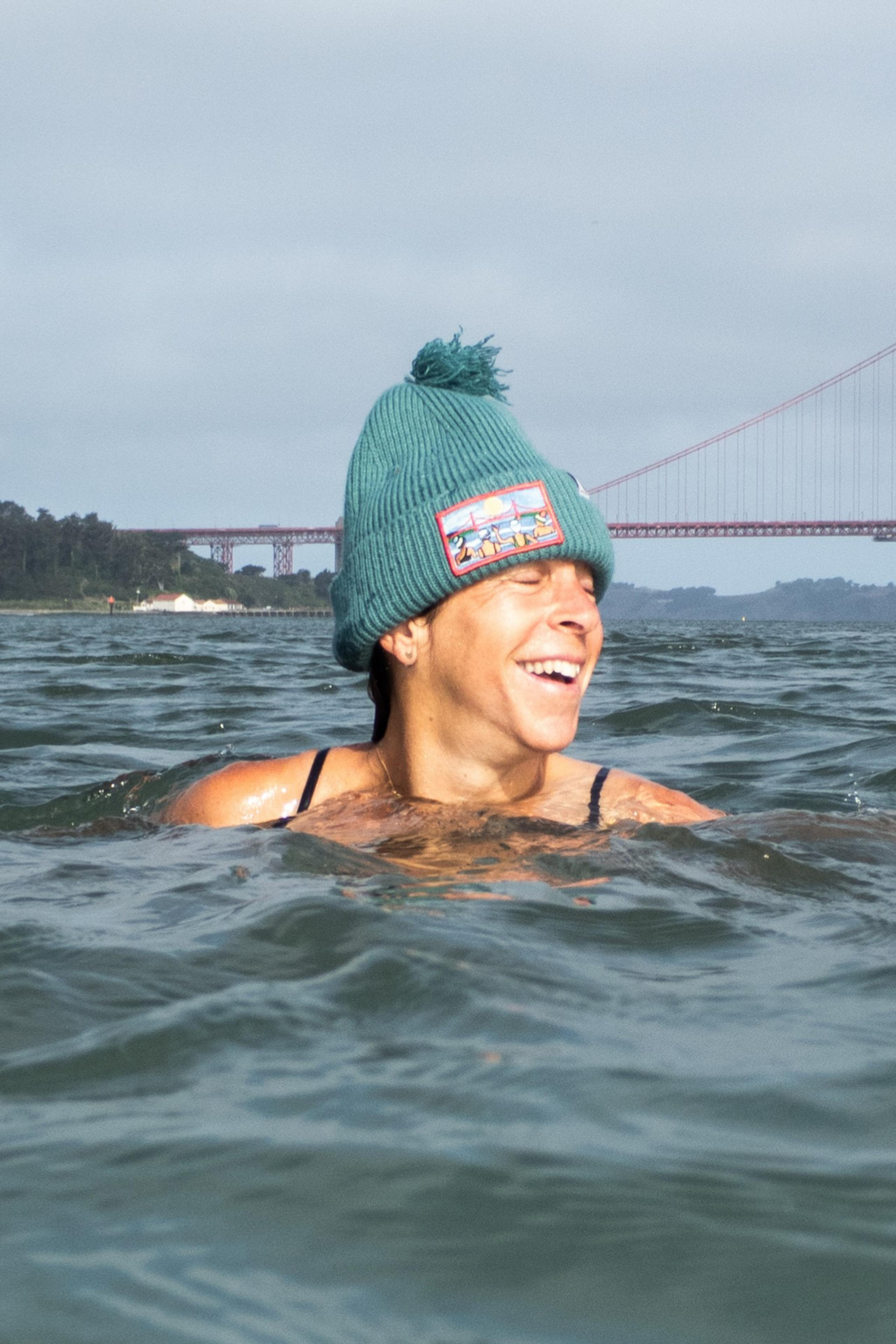 A person in a beanie with a patch smiles joyfully while swimming in the water, with a large suspension bridge in the background under a cloudy sky.