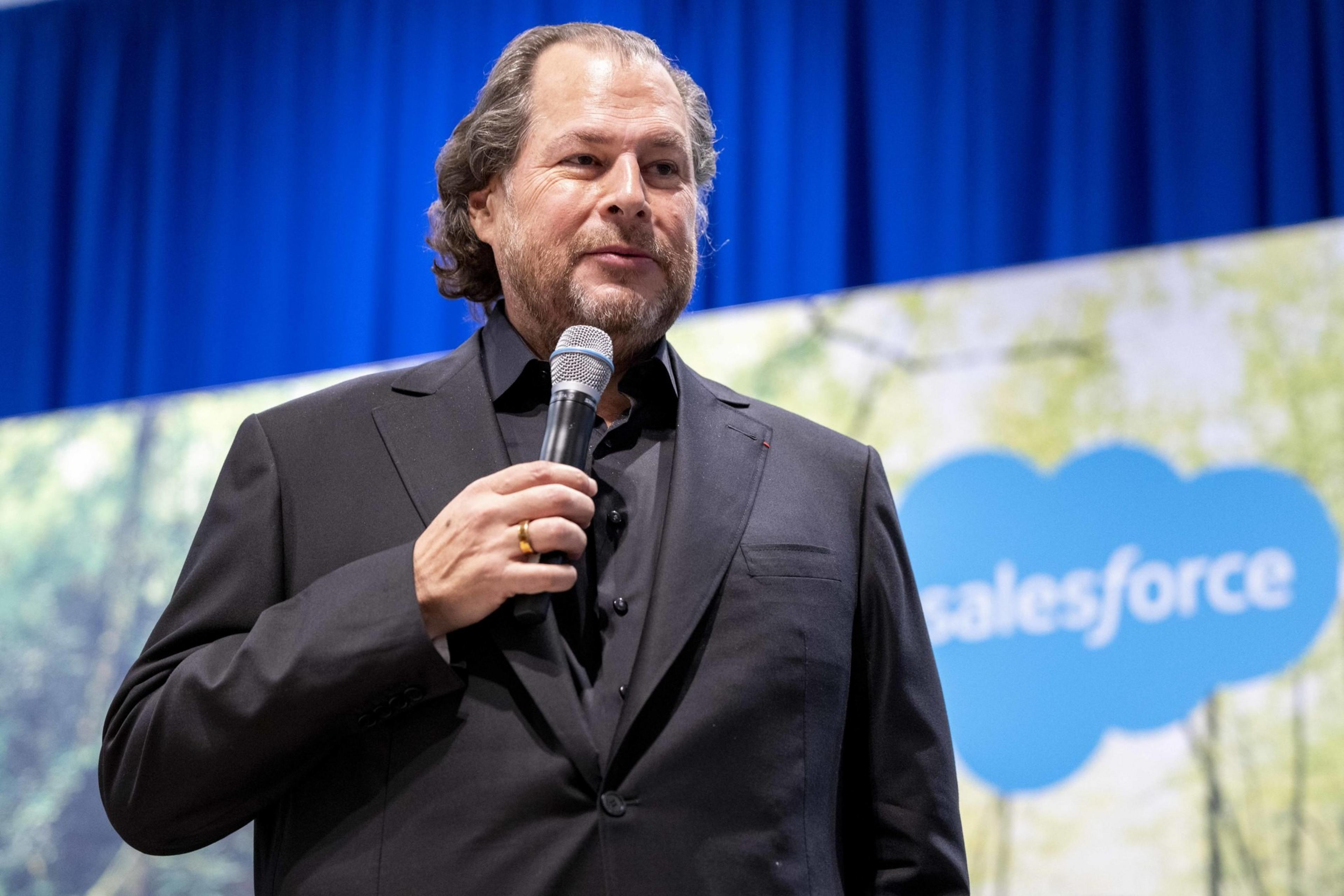 A man in a black suit is speaking into a microphone on stage. Behind him is a blurred blue and green background featuring a cloud-shaped Salesforce logo.