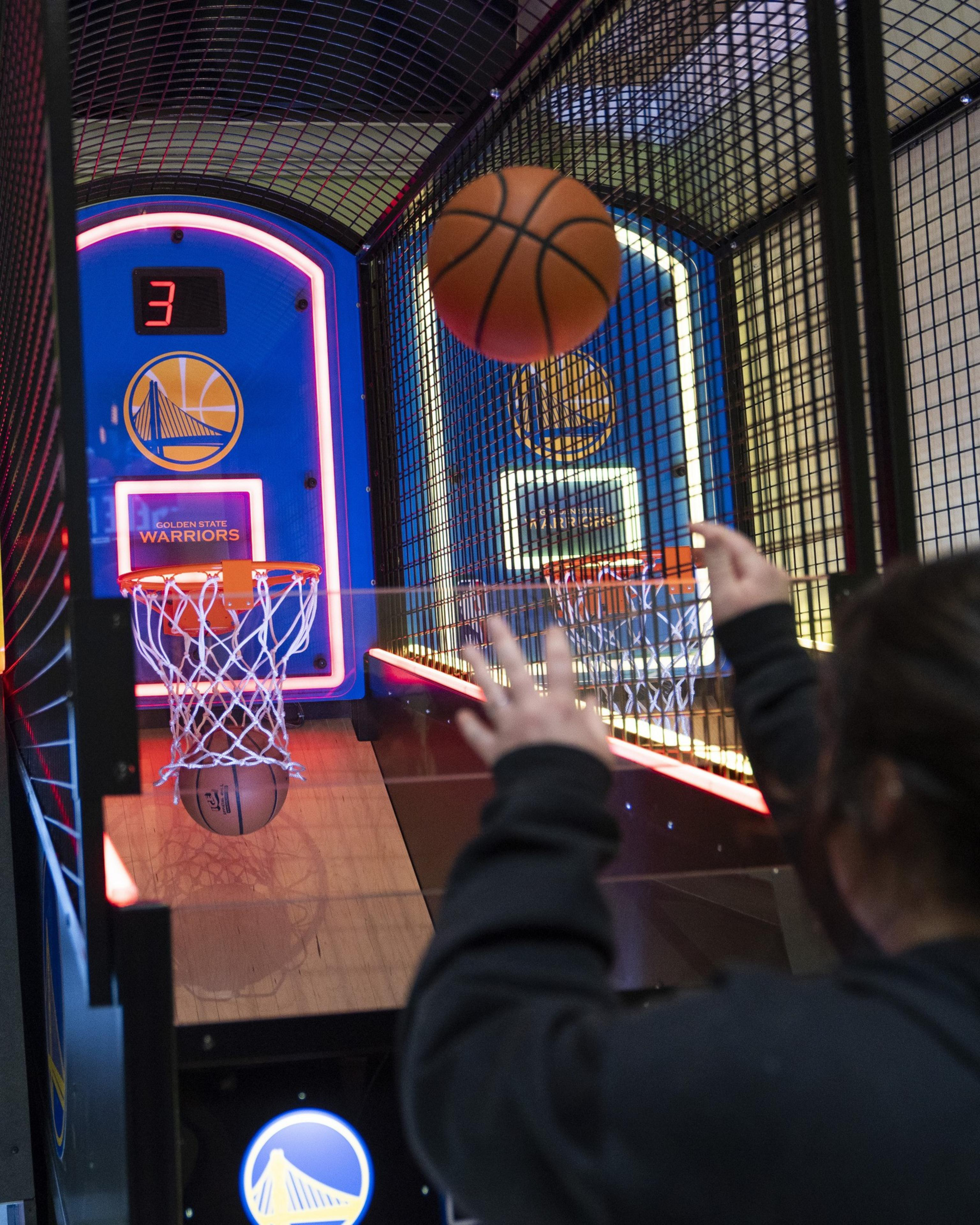 A person is playing arcade basketball, aiming at a hoop. The score display shows the number 3, and the setup is decorated with Golden State Warriors branding.