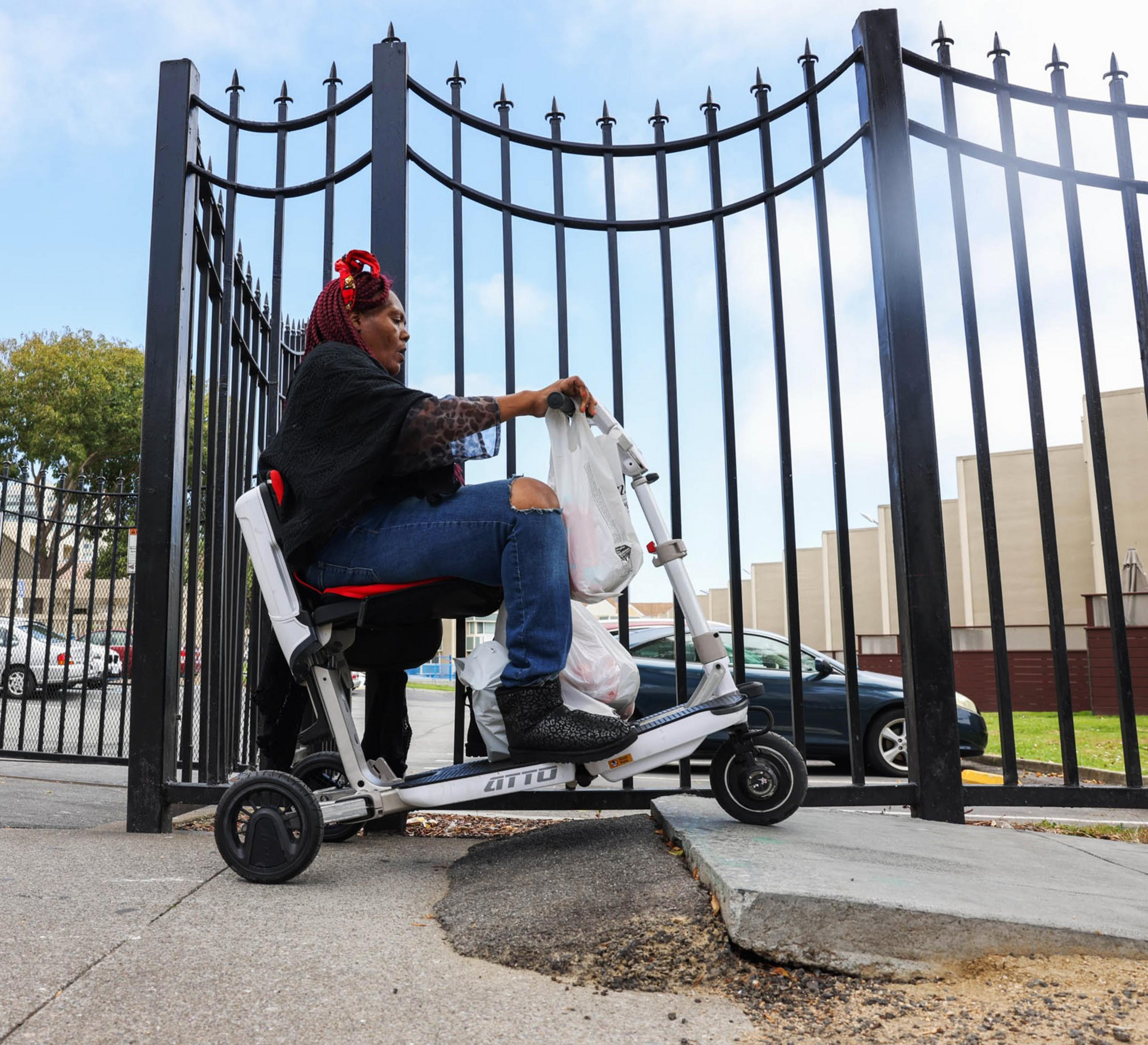 A woman with red hair braids rides a motorized scooter, carrying bags, and navigates up a curb ramp. She's next to a tall black iron fence with a cityscape in the background.