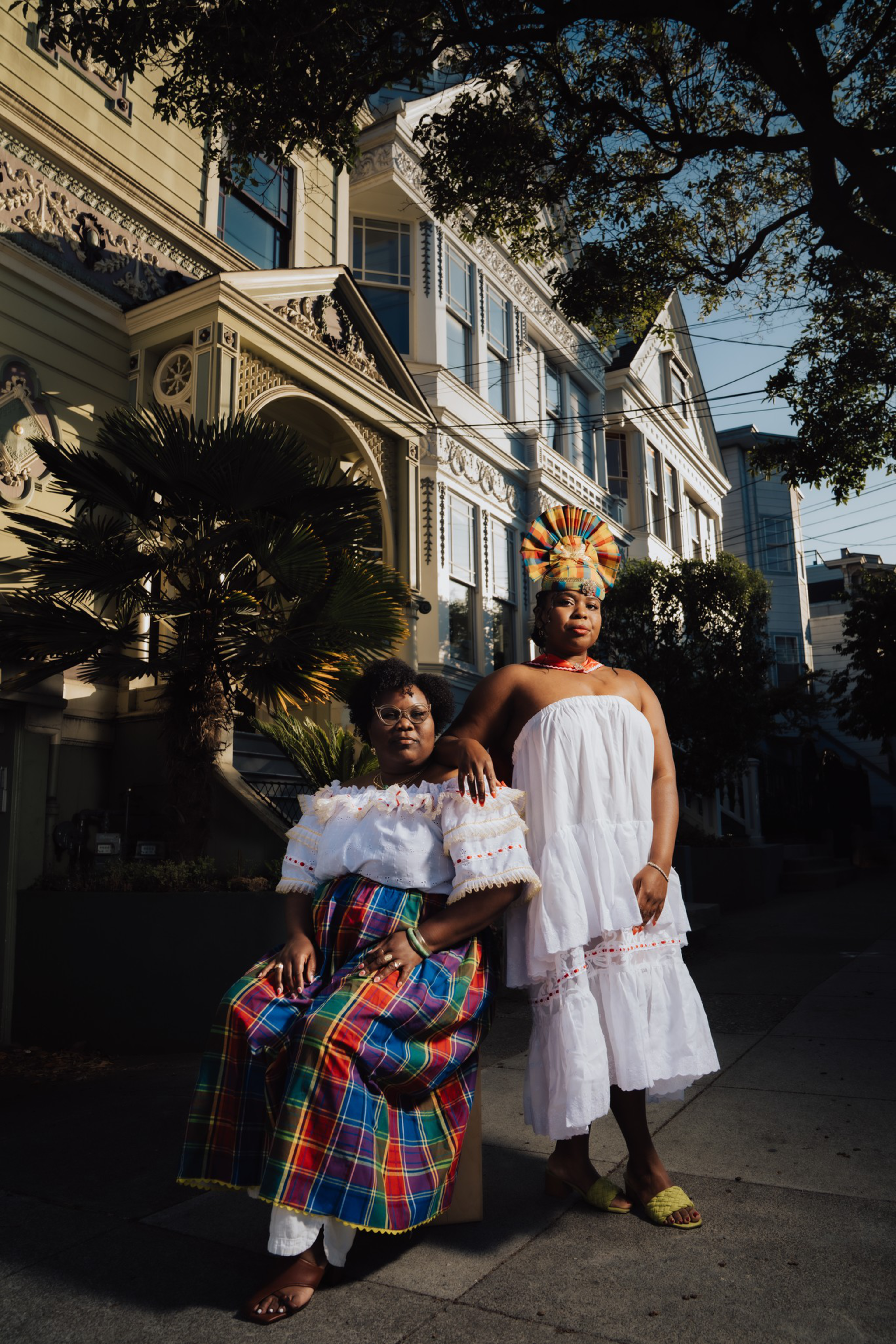 Two women pose confidently on a sunlit sidewalk in front of ornate Victorian buildings; one wears a vibrant plaid skirt and off-shoulder top, the other a white dress and colorful headwrap.