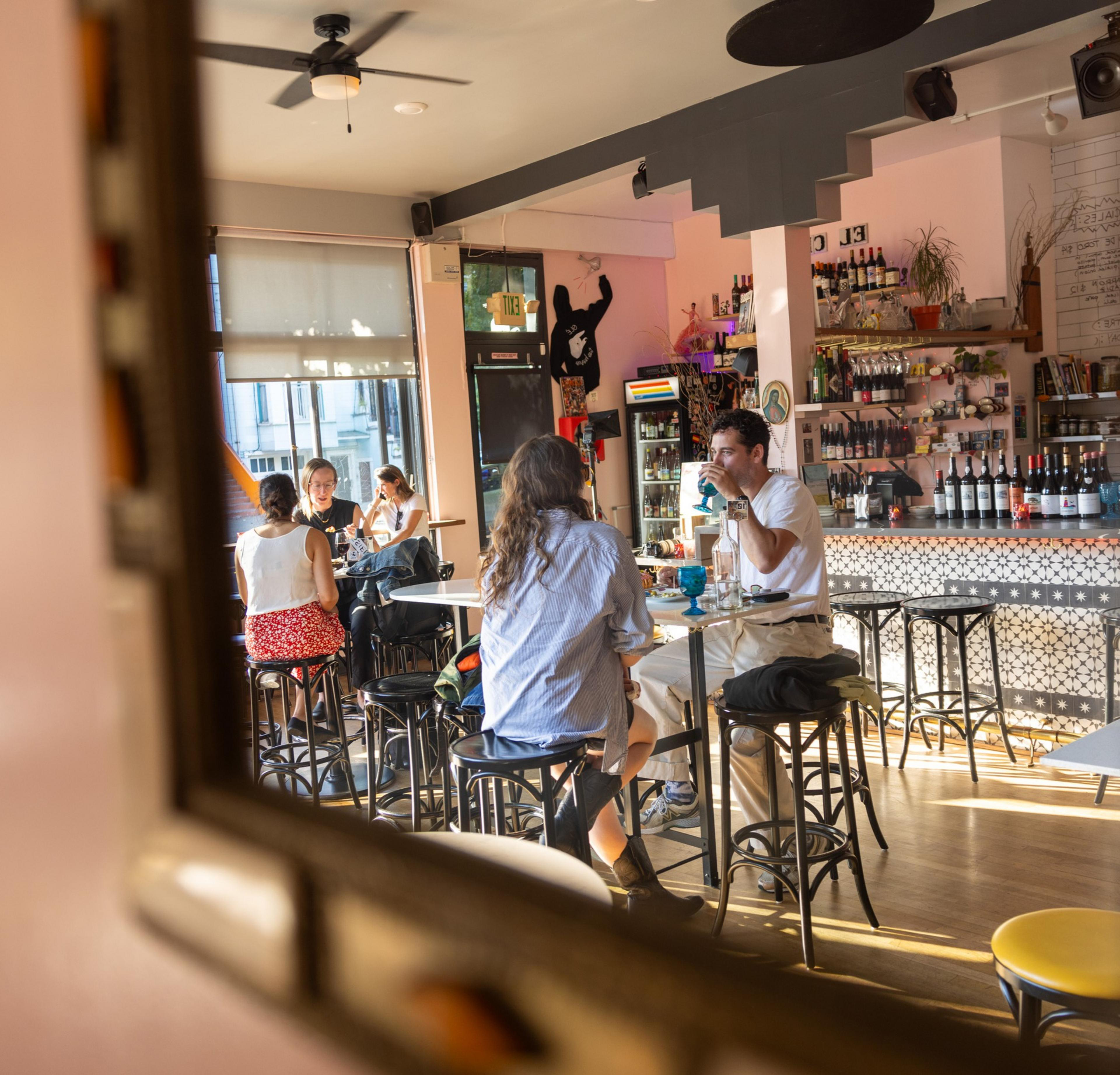 A cozy café with several people sitting at small tables, conversing and enjoying drinks. The décor includes a well-stocked bar, warm lighting, and a playful mural.