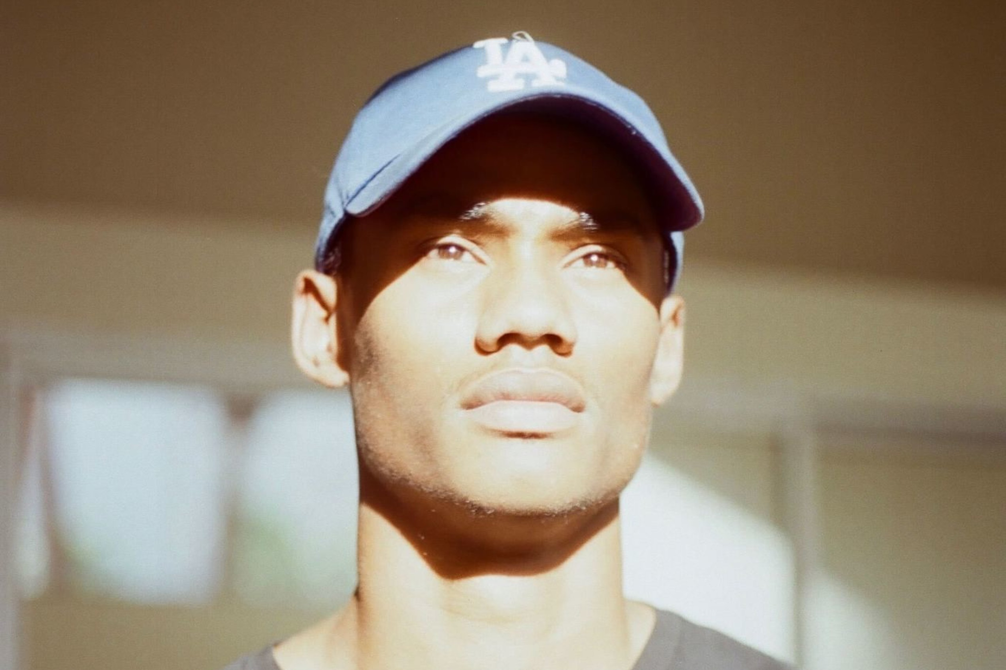 A man wearing a blue LA cap and black t-shirt looks upward with sunlight casting shadows on his face and shirt.