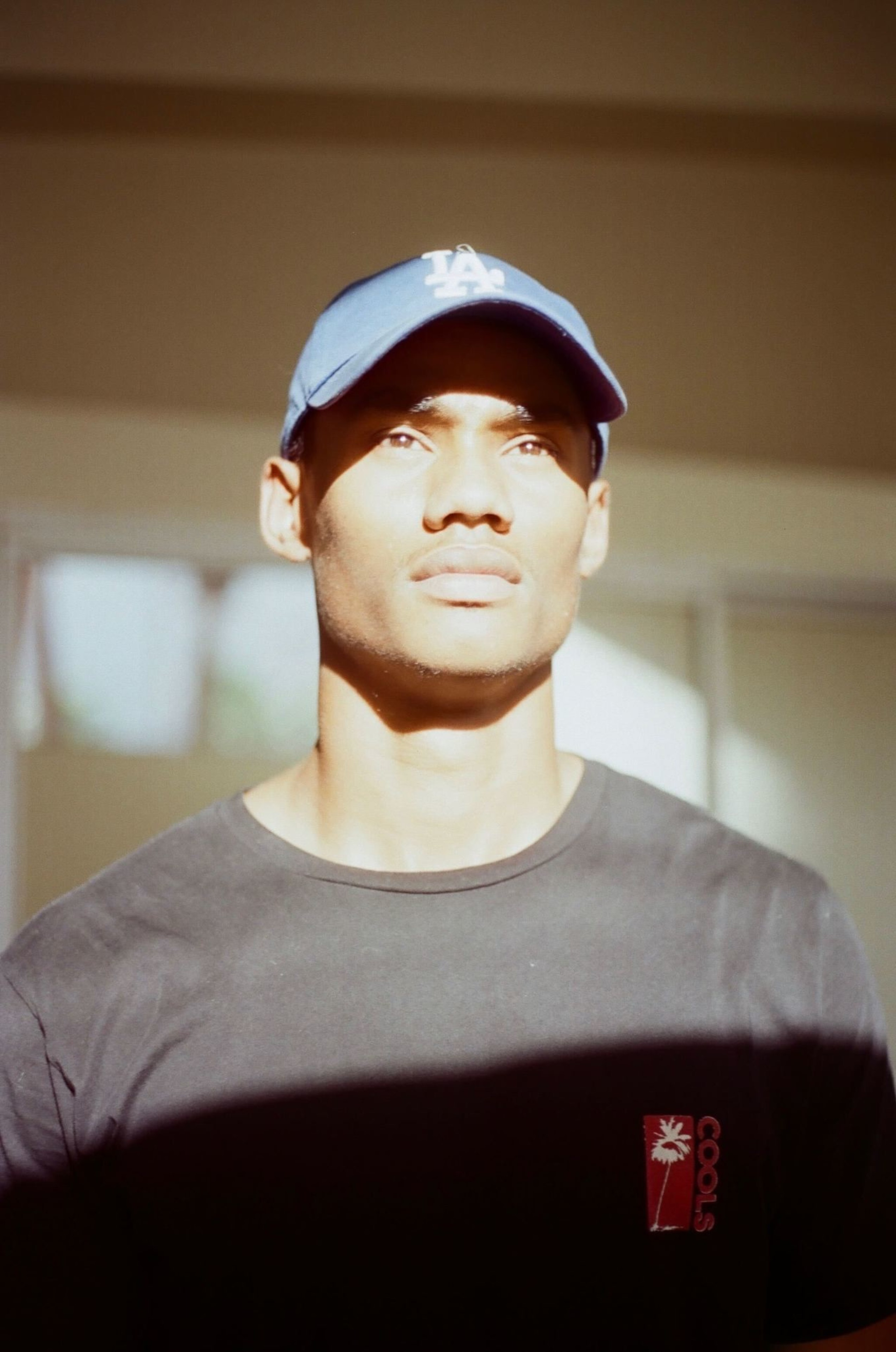A man wearing a blue LA cap and black t-shirt looks upward with sunlight casting shadows on his face and shirt.