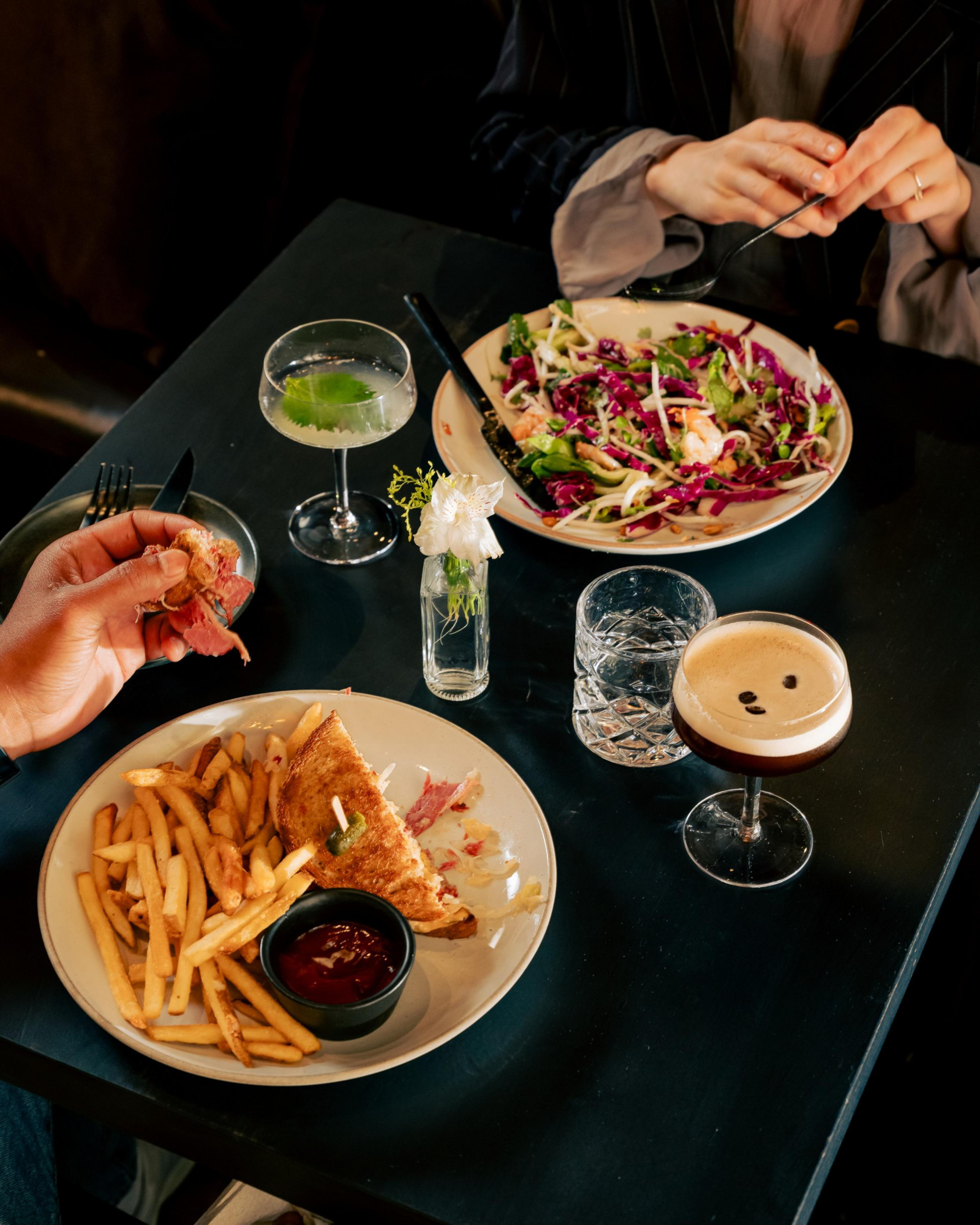 A table set with a salad, sandwich, fries, cocktails, and two diners. One holds a sandwich, another adjusts utensils. There's a small flower vase on the table.
