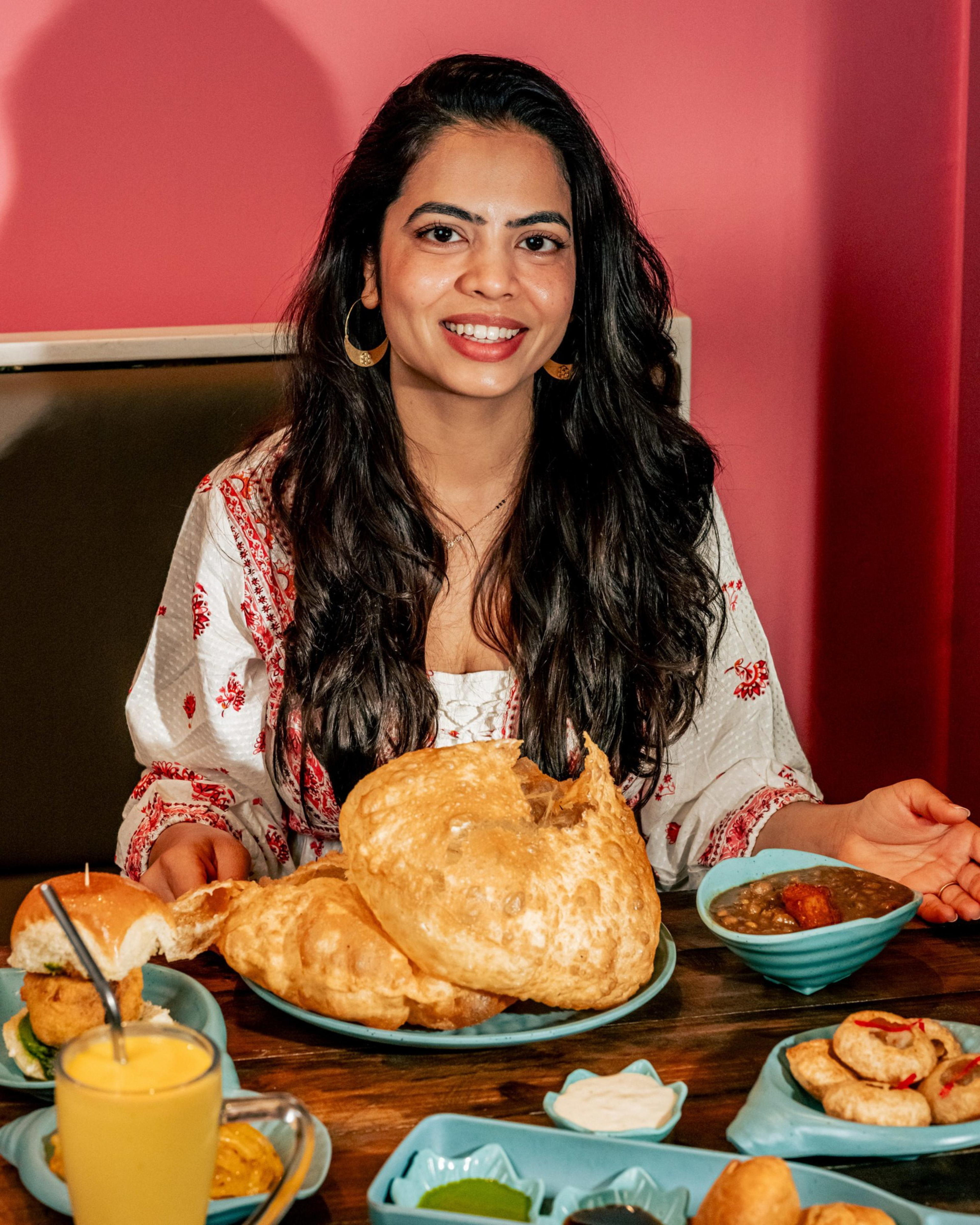 A woman with long dark hair smiles while sitting at a table filled with Indian dishes, including puris, curried lentils, snacks, and a glass of mango drink.