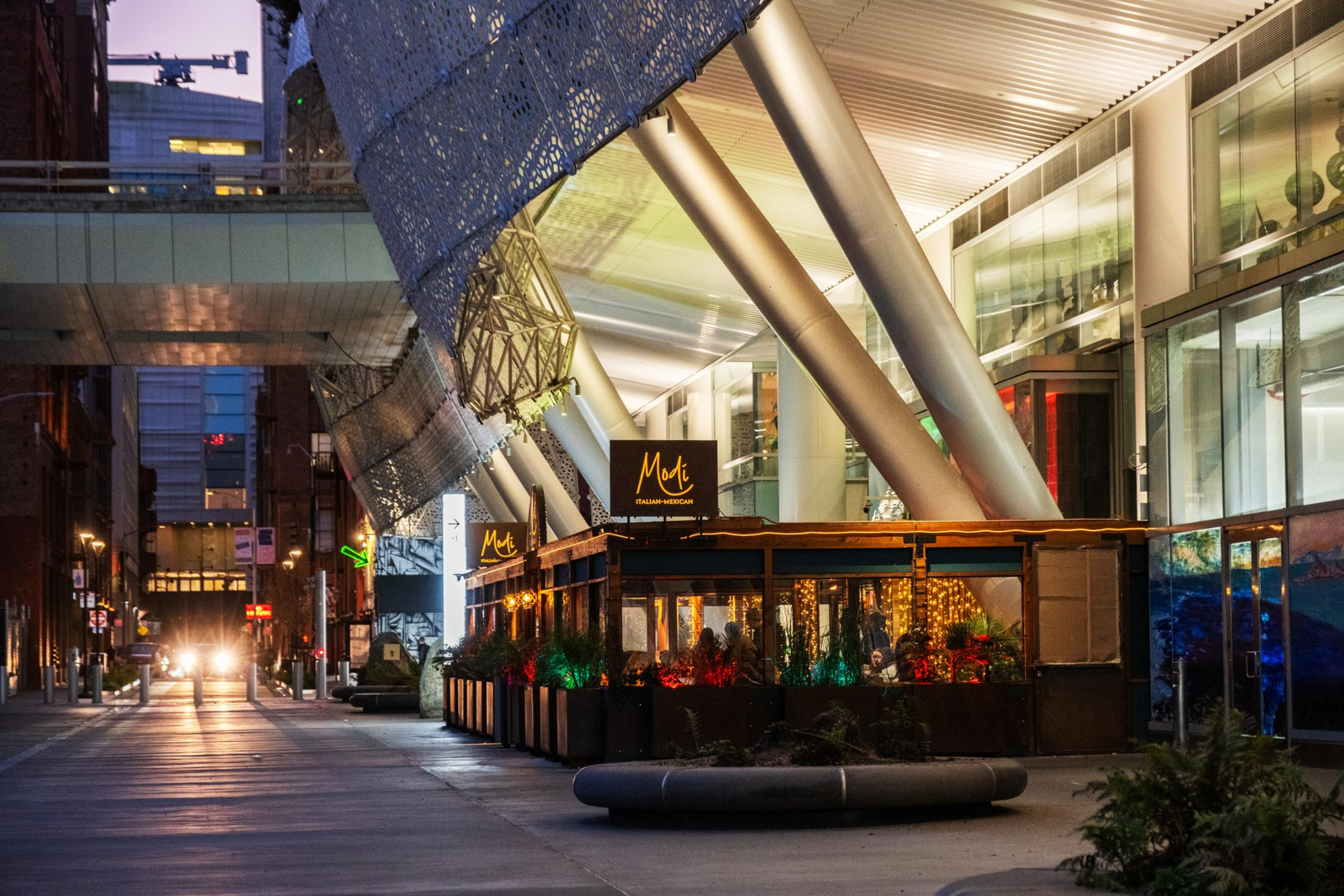The image shows a modern urban area with a building featuring large metal pillars. A restaurant with the sign "Modi" is in the foreground, illuminated by warm lights.