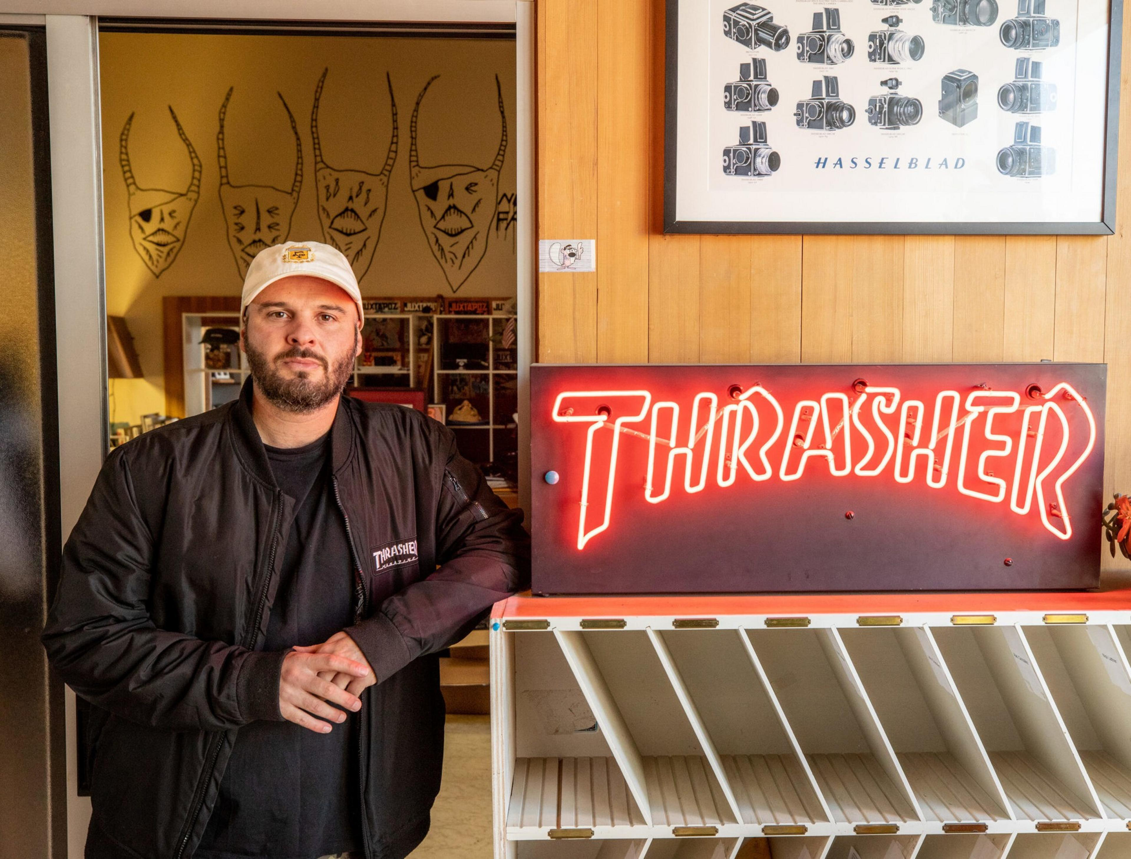 A man wearing a black Thrasher jacket and white cap leans against a wall with a red neon "THRASHER" sign and a framed Hasselblad camera poster above it.