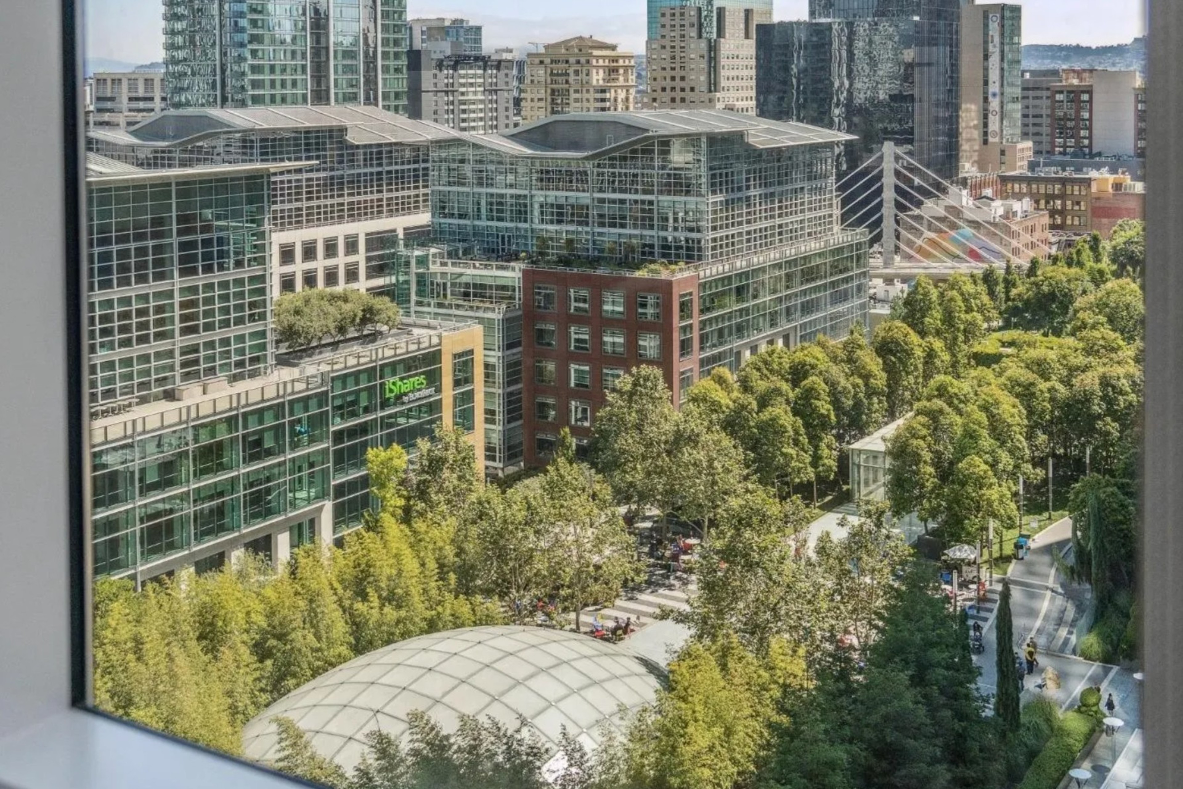 A cityscape featuring modern glass buildings and a park with dense trees. Within the park, a glass dome structure is seen, and a bridge is visible in the background.