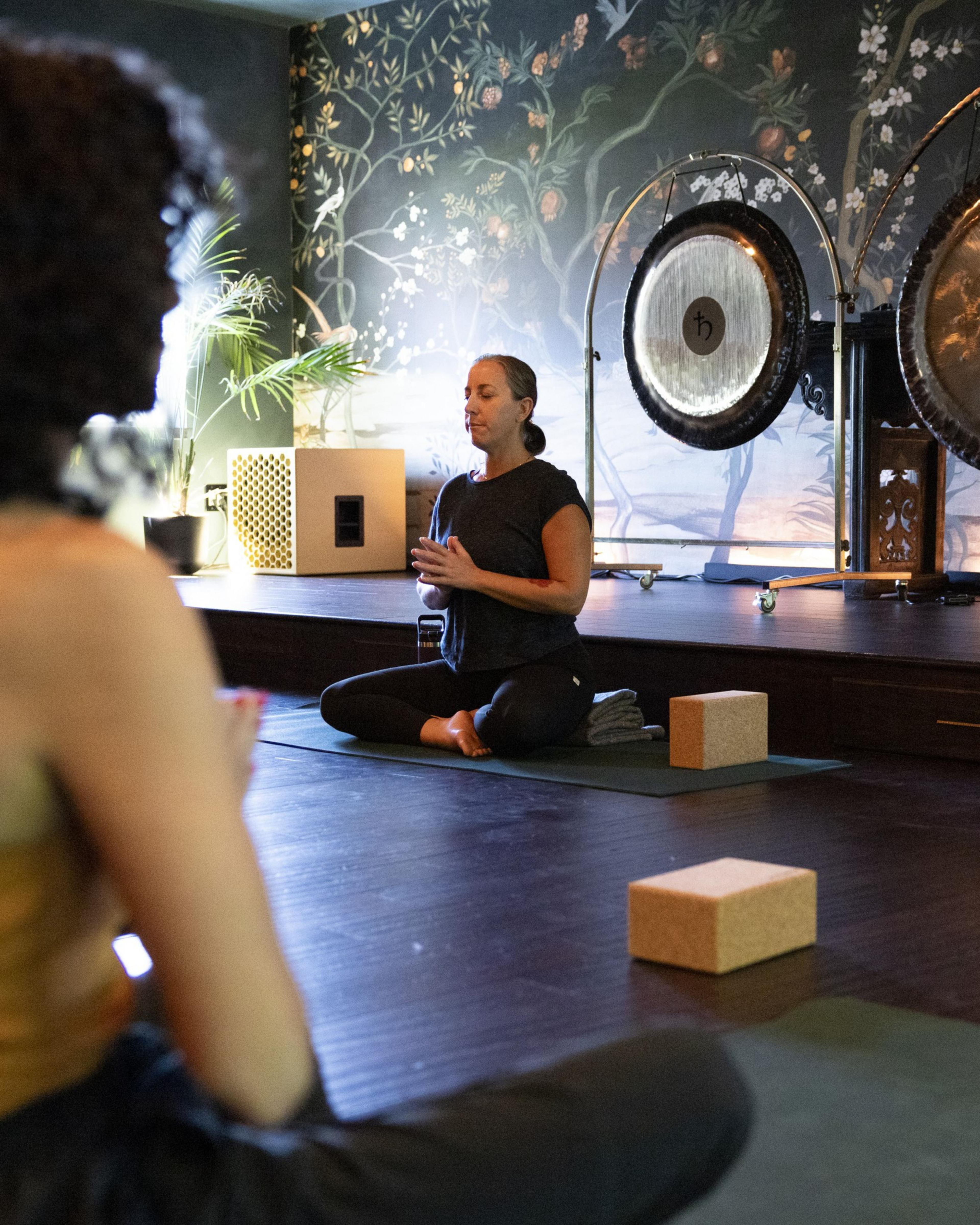 A woman sits cross-legged in a meditation pose on a yoga mat, with another person in the foreground. The room has floral wallpaper and large gongs.