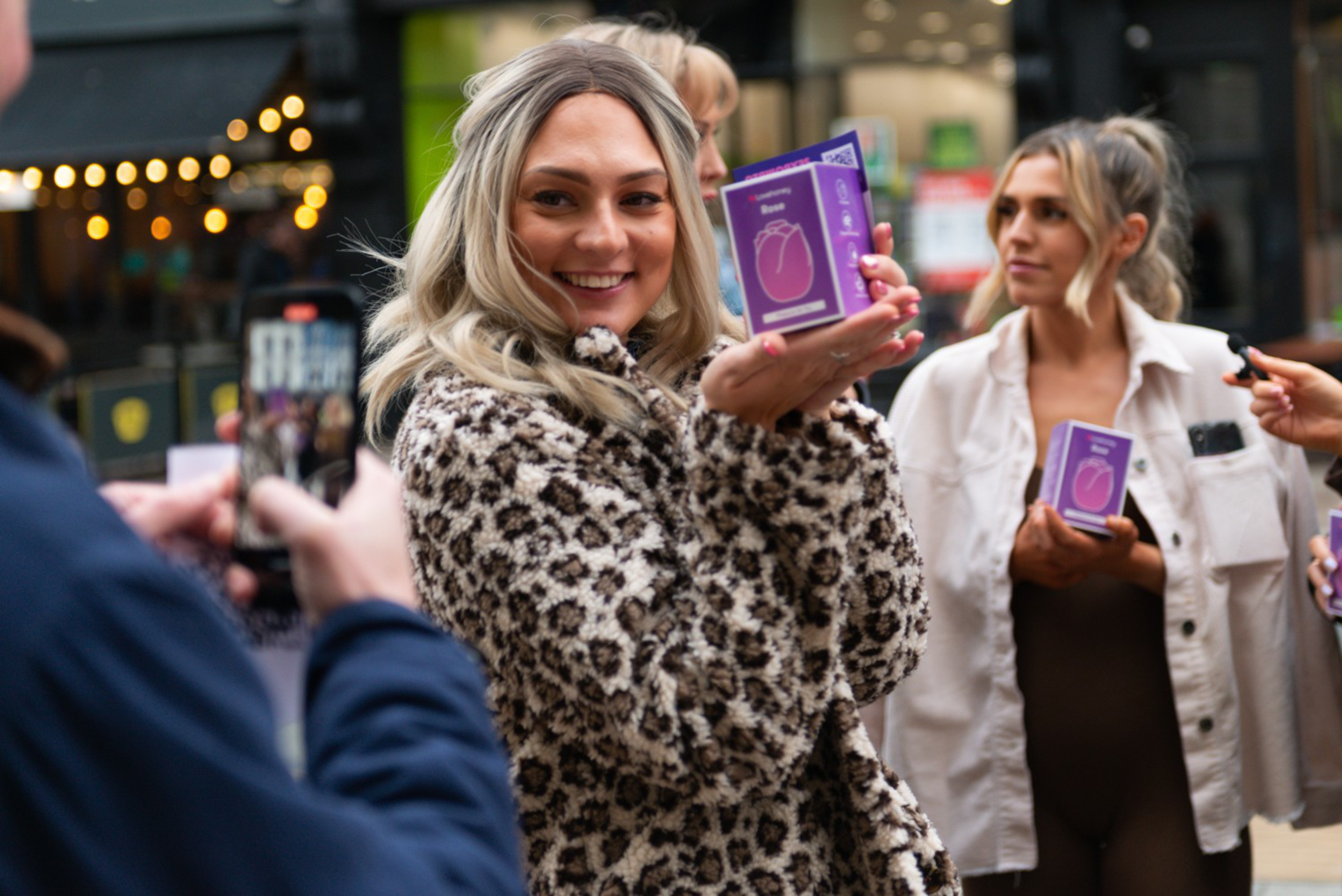 A woman in a leopard print coat smiles while holding a purple boxed product. Another person in white holds a similar box. A person nearby takes a photo.